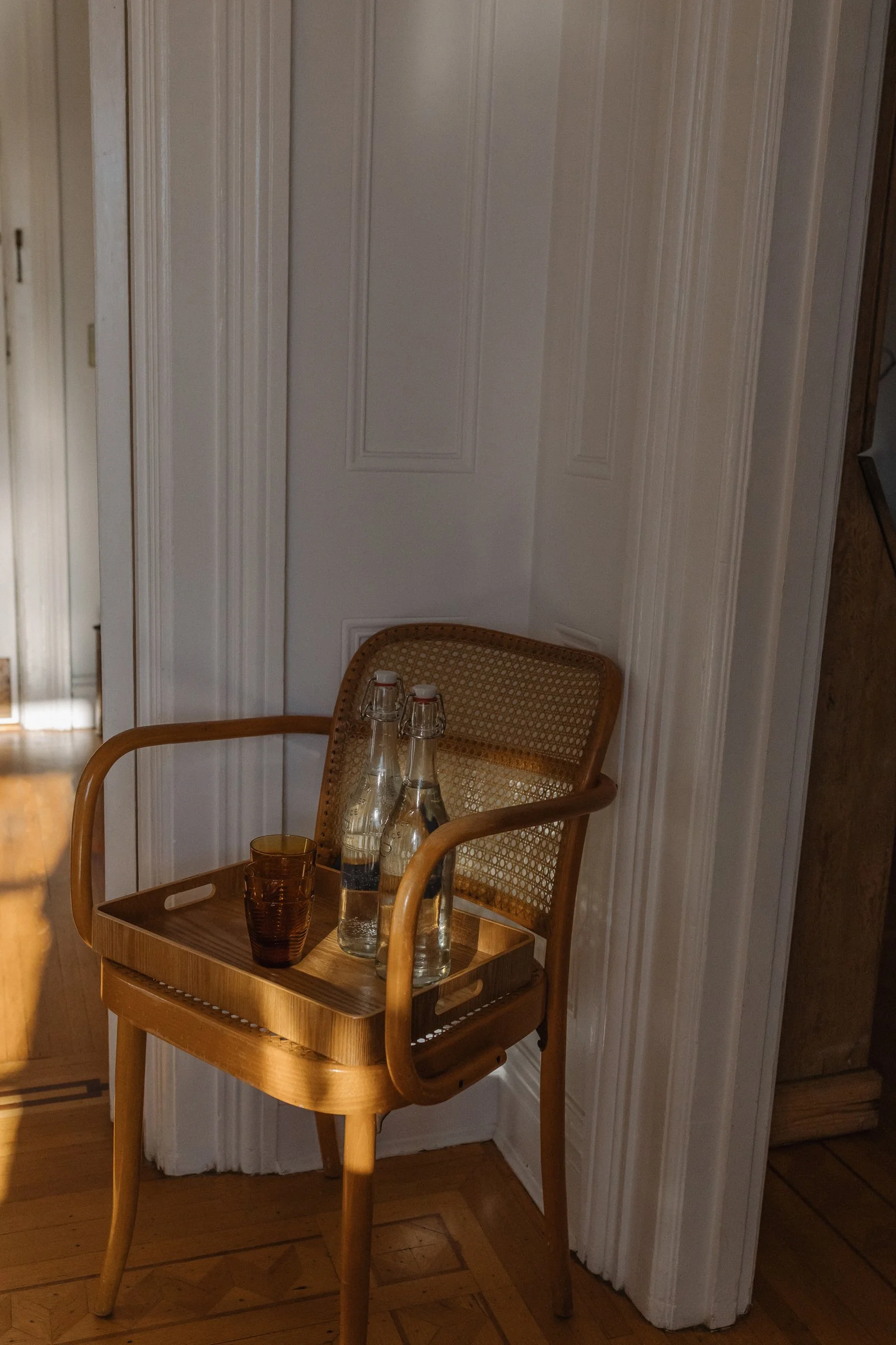 A wooden chair with a tray holding two glass bottles of water and a brown glass in a sunlit room with white paneled walls and wooden flooring.