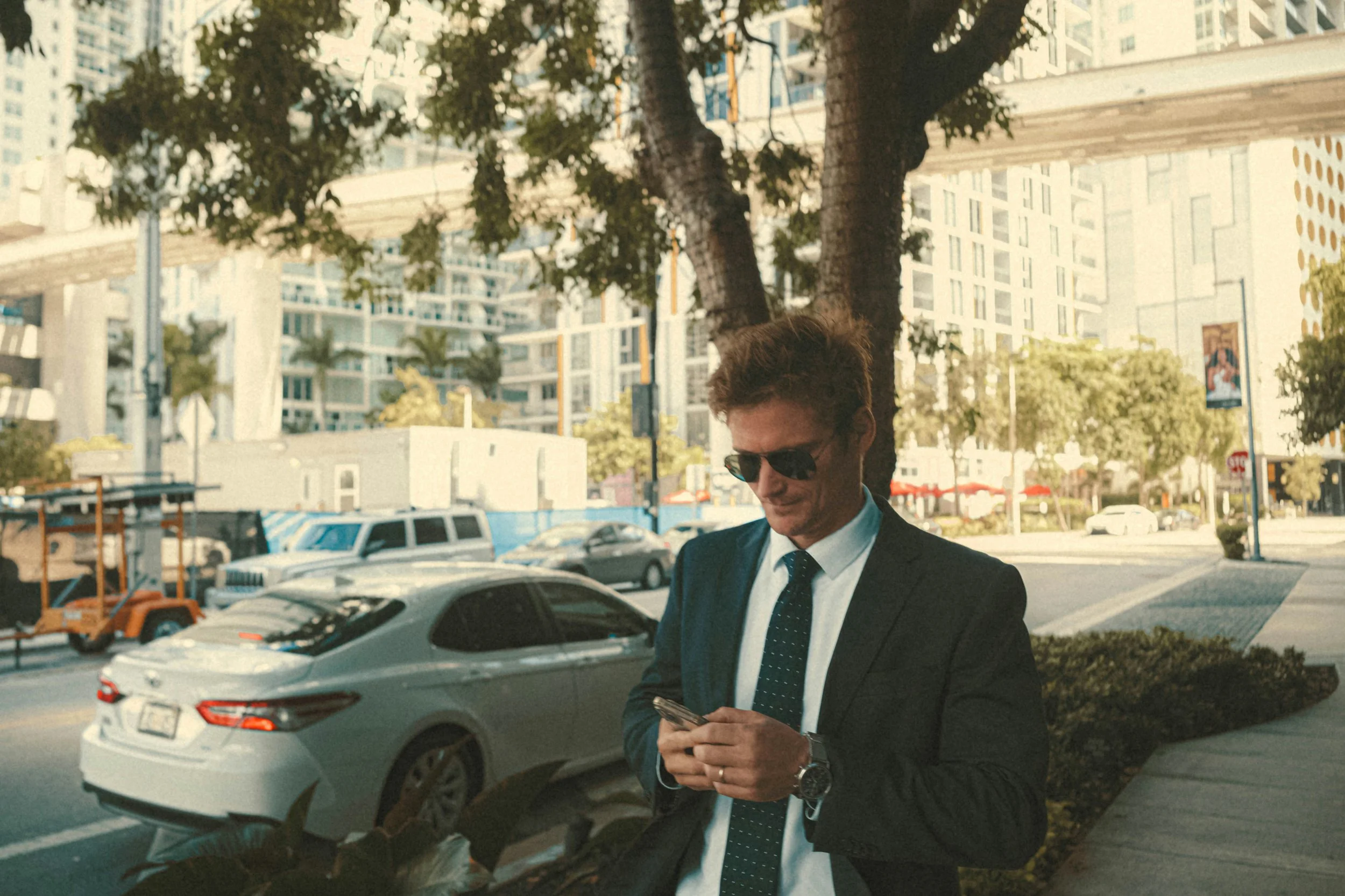 A man in a business suit and sunglasses stands on a sidewalk, looking at his phone, with cars and tall buildings in the background.