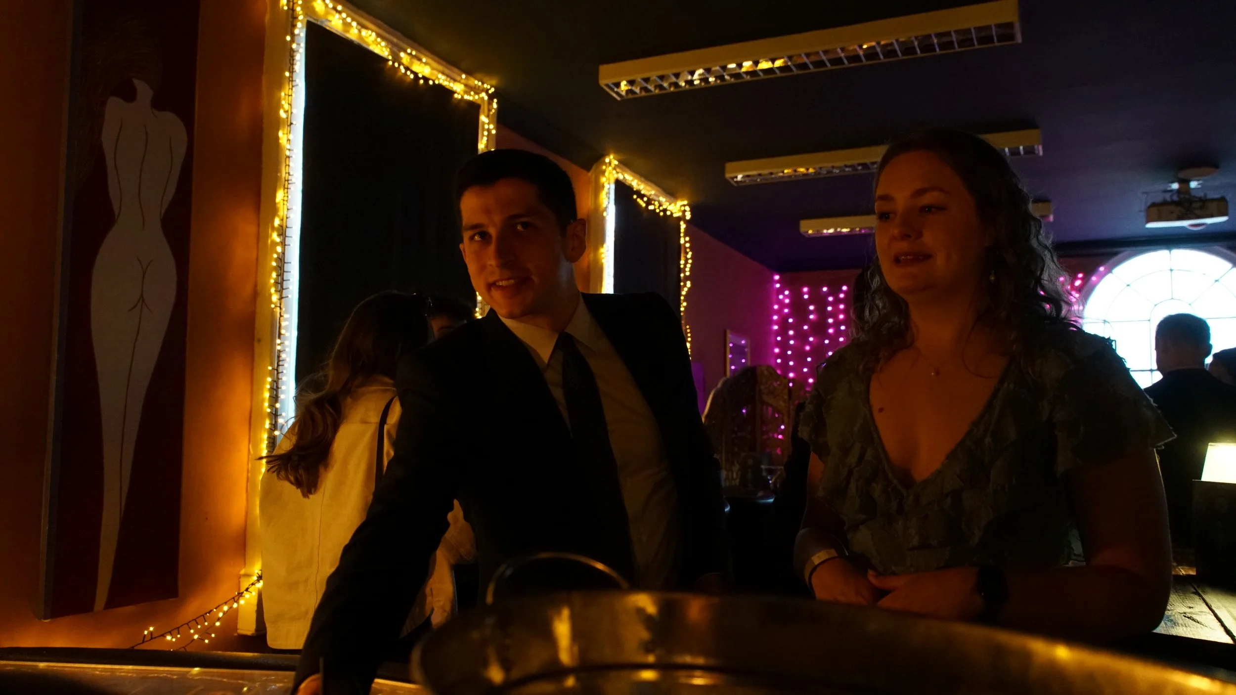 A man in a suit and a woman are standing at a bar in a dimly lit room decorated with string lights and colorful lights. The man is smiling, and the woman looks slightly emotional.