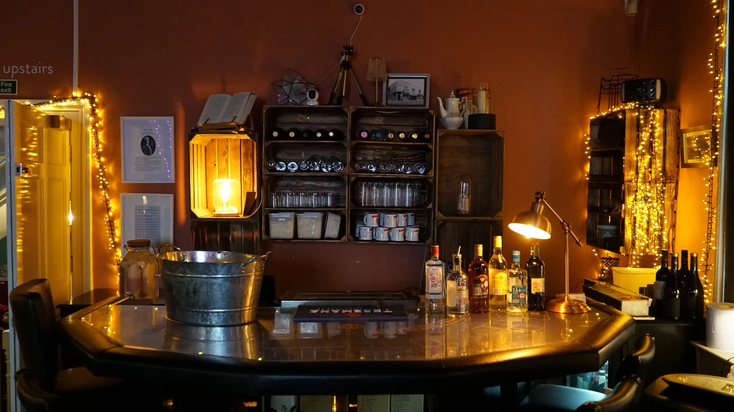 A cozy bar scene with dim lighting, featuring a wooden bar counter with various bottles of alcohol, a small lamp, and decorative string lights on the walls. There are shelves with glassware, a bucket on the counter, and framed pictures on the wall.