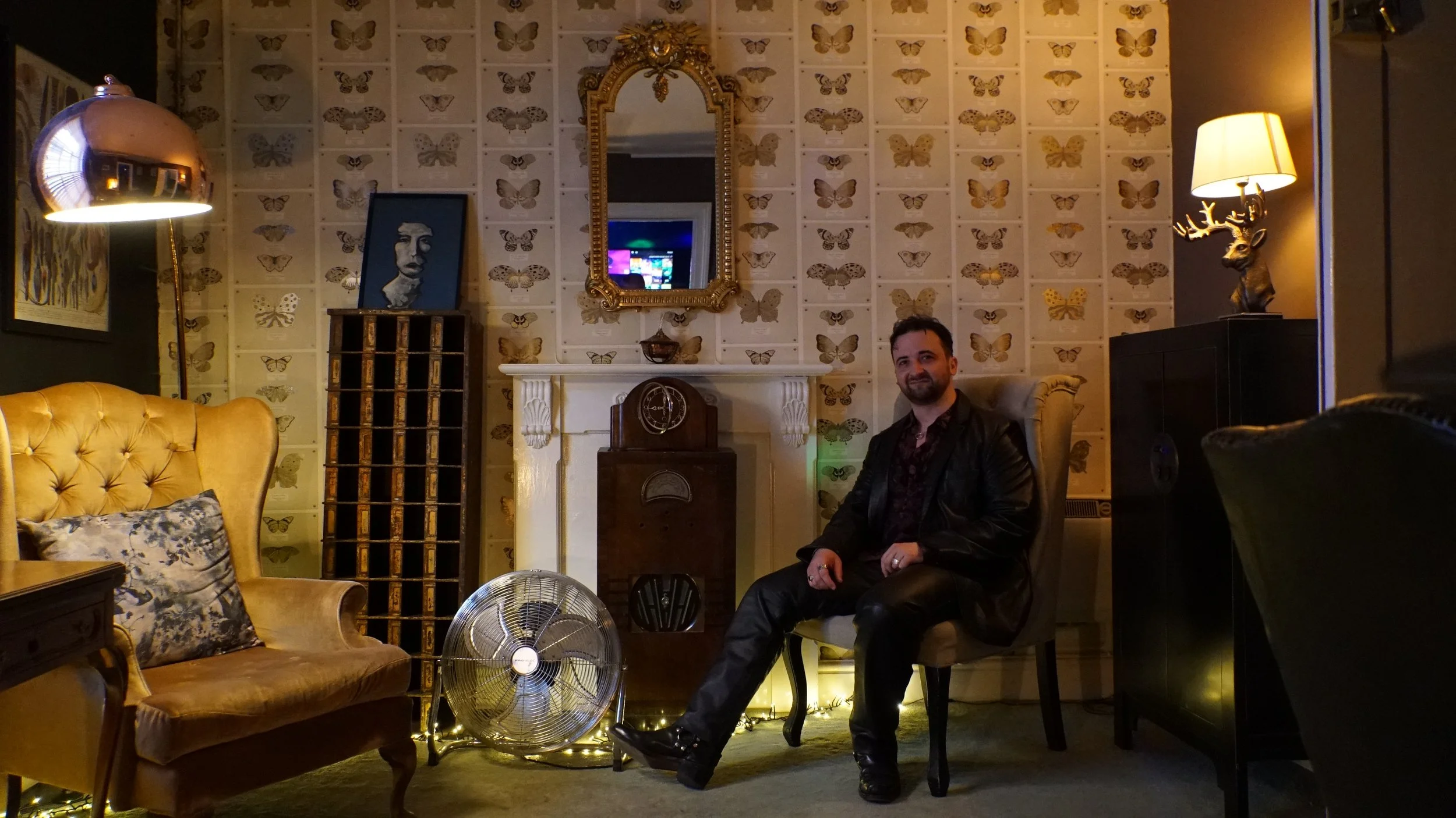 A man sitting on a chair in a warmly decorated living room with butterfly wallpaper, a vintage radio, a yellow tufted sofa, a fan, and decorative lamps.