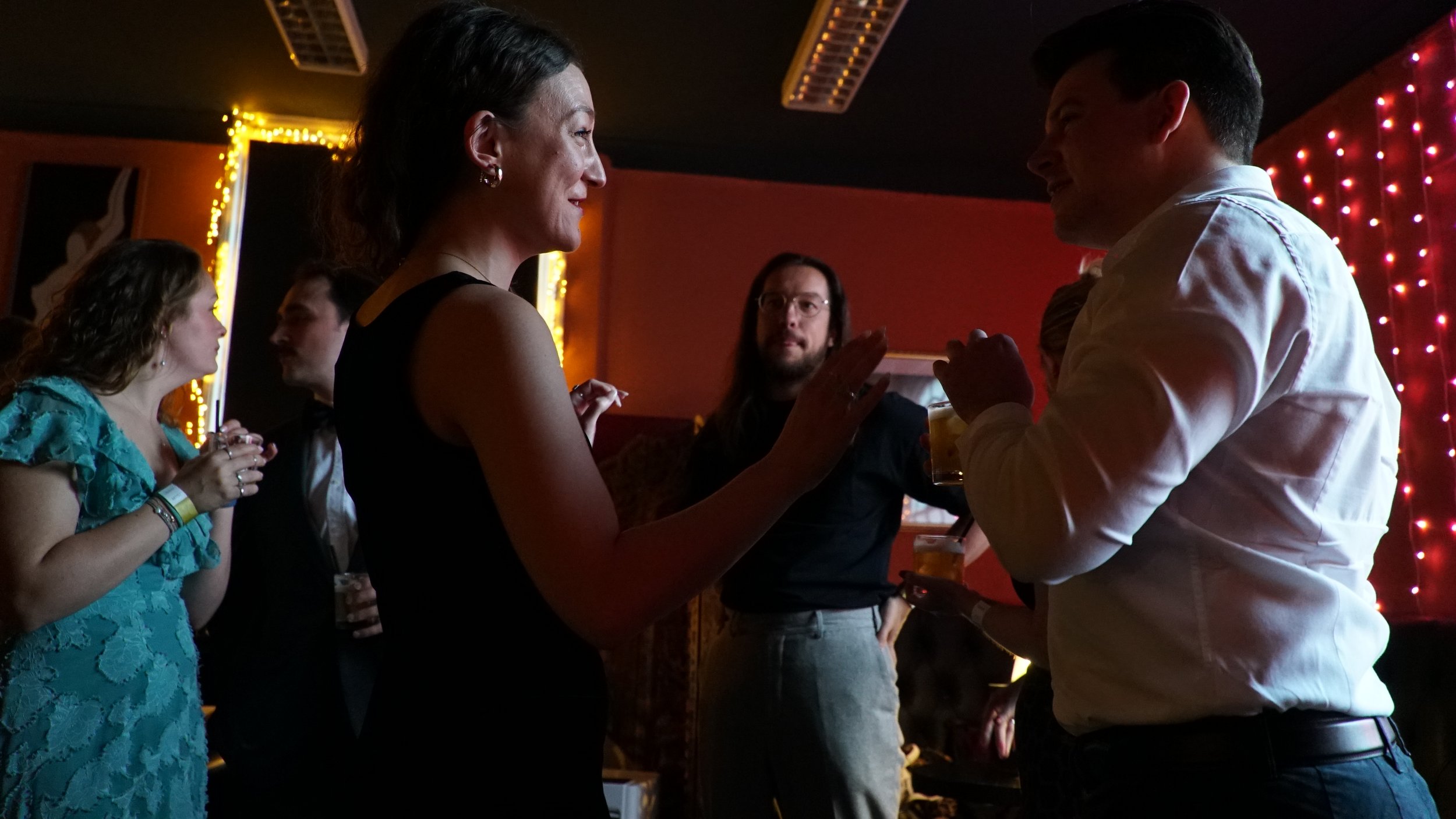 Group of people socializing indoors at a dimly lit party or bar, with some holding drinks and engaging in conversation.