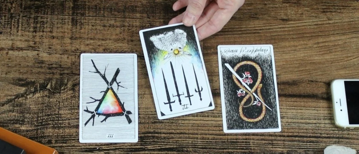 Three Tarot cards laid out on a wooden table, with a person holding one card. The cards feature symbolic illustrations, including a triangle with branches, five swords, and a snake intertwined with flowers and a sword.