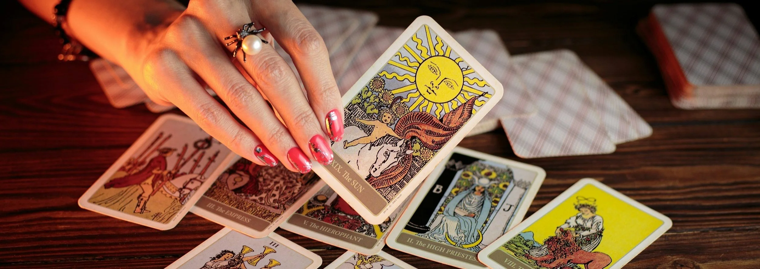 A hand with decorative red nail polish and jewelry reaches over tarot cards spread on a wooden surface, including the Sun card with a smiling sun and face.