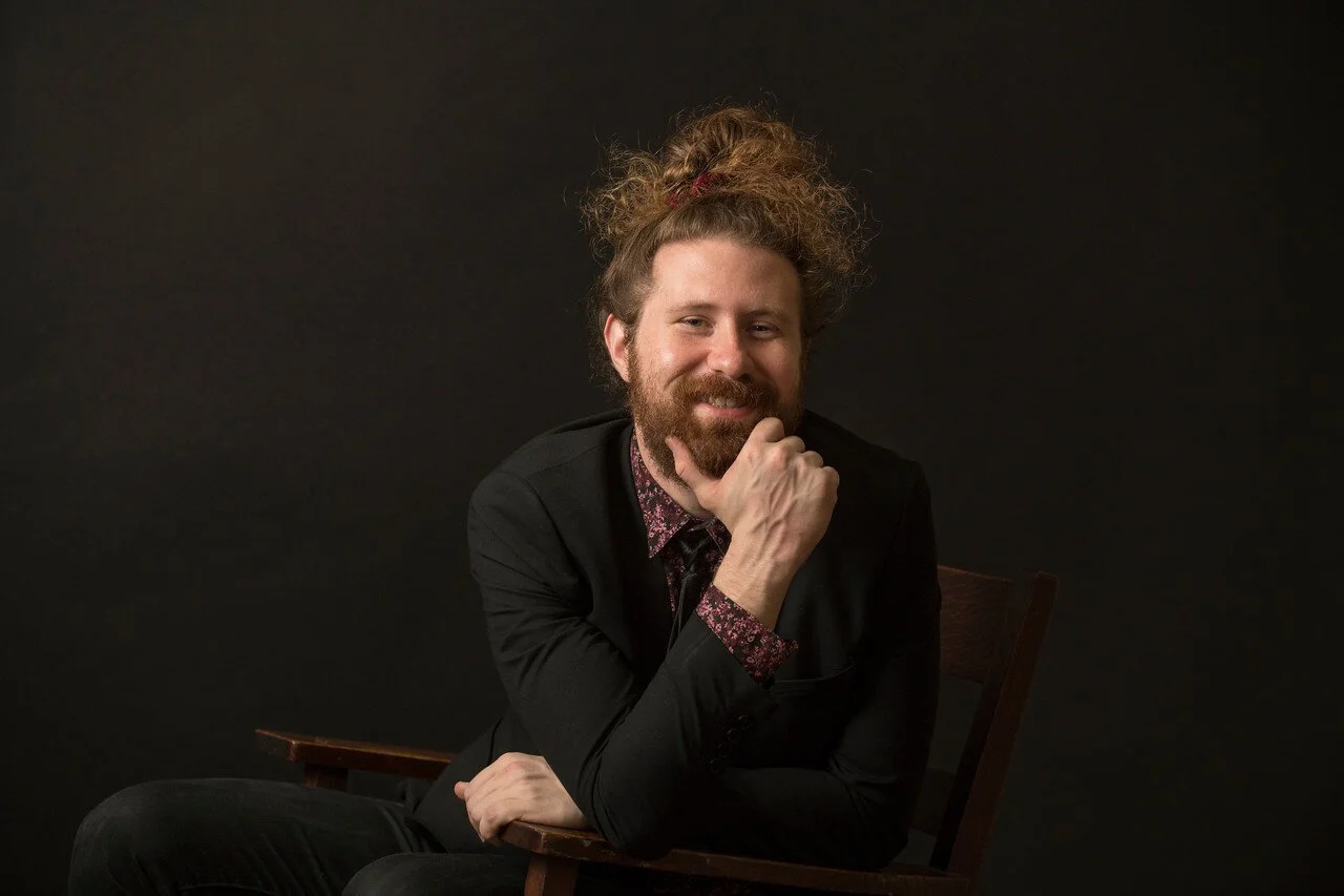 Casey Abrams, smiling, wearing a black jacket and floral shirt, seated against a dark background.