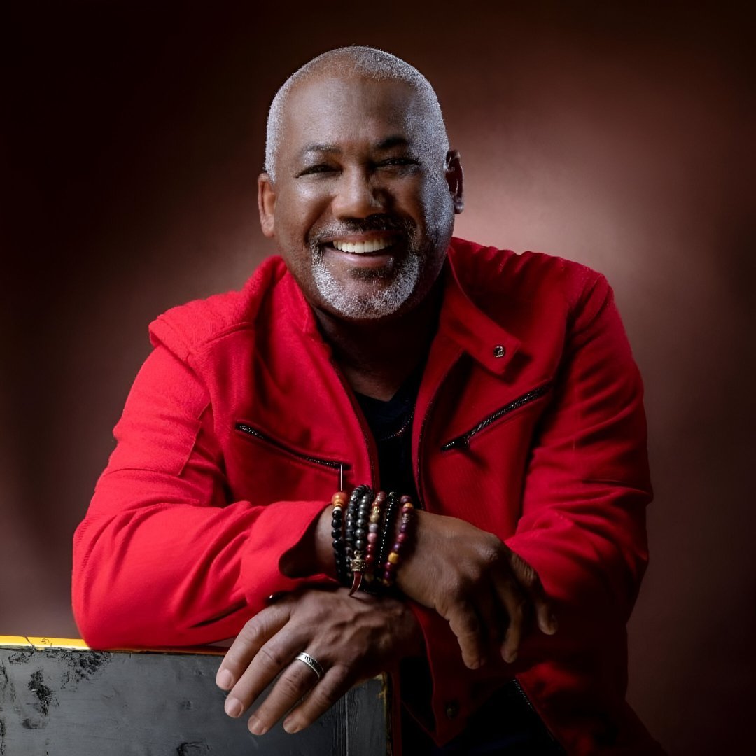 Jonathan Butler wearing a red jacket, sitting with arms crossed and decorated with multiple bracelets, against a dark, blurred background.