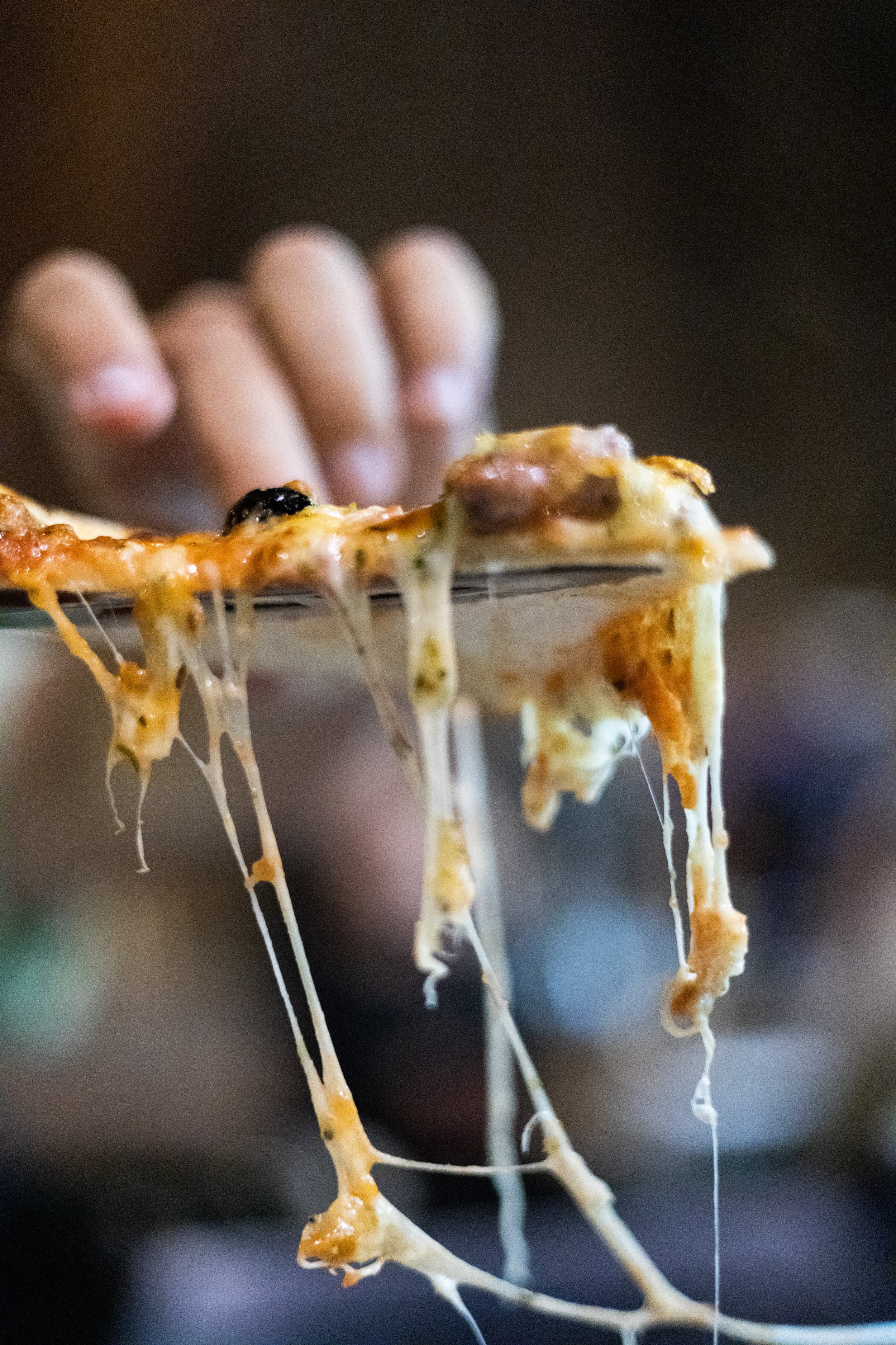 Close-up of a slice of Marseille's pizza with melted cheese hanging off the edge, held by a hand in the background.