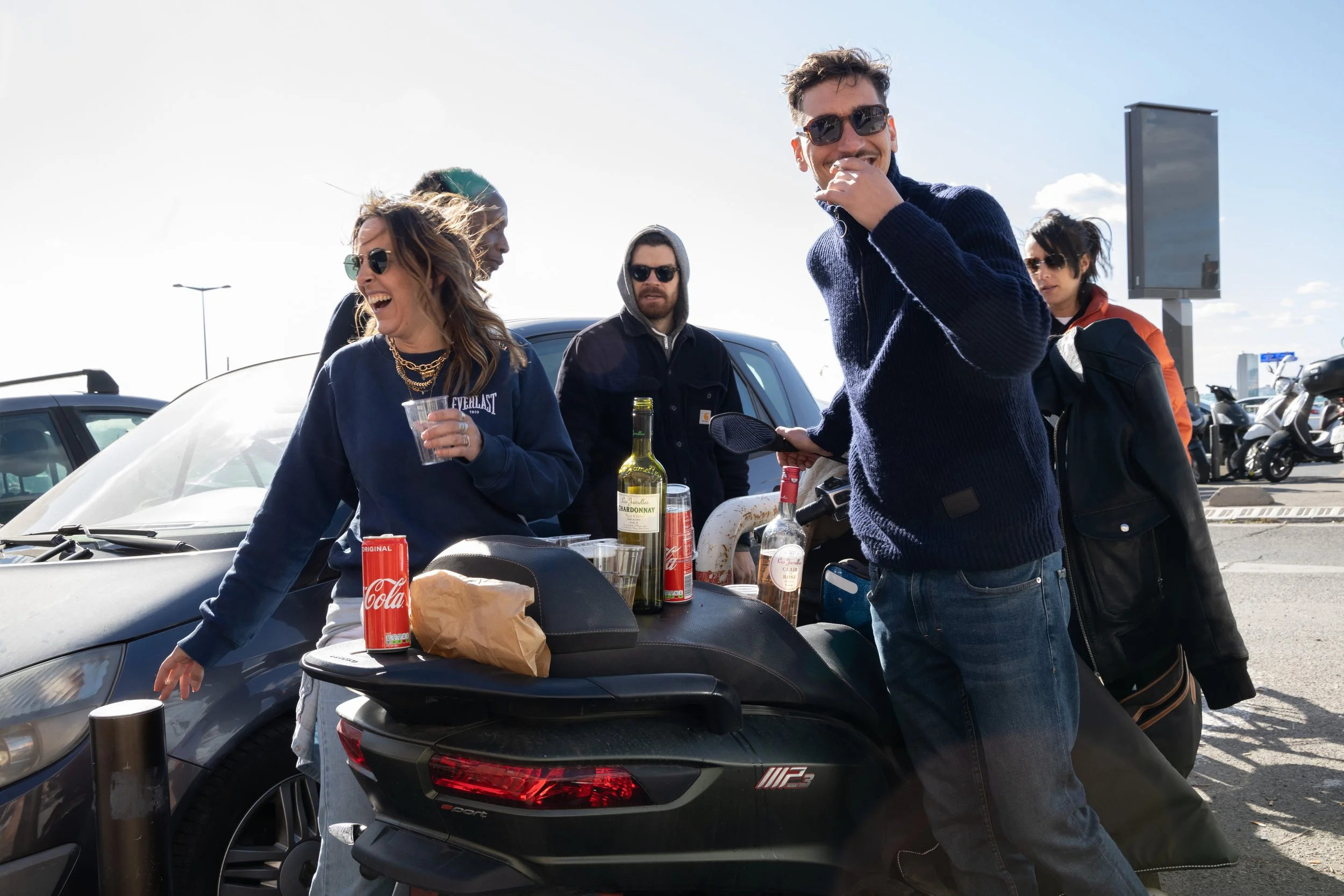 Group of people socializing at an outdoor parking lot in Marseille, with drinks and snacks, sunny weather, some wearing sunglasses, and a scooter in the foreground.