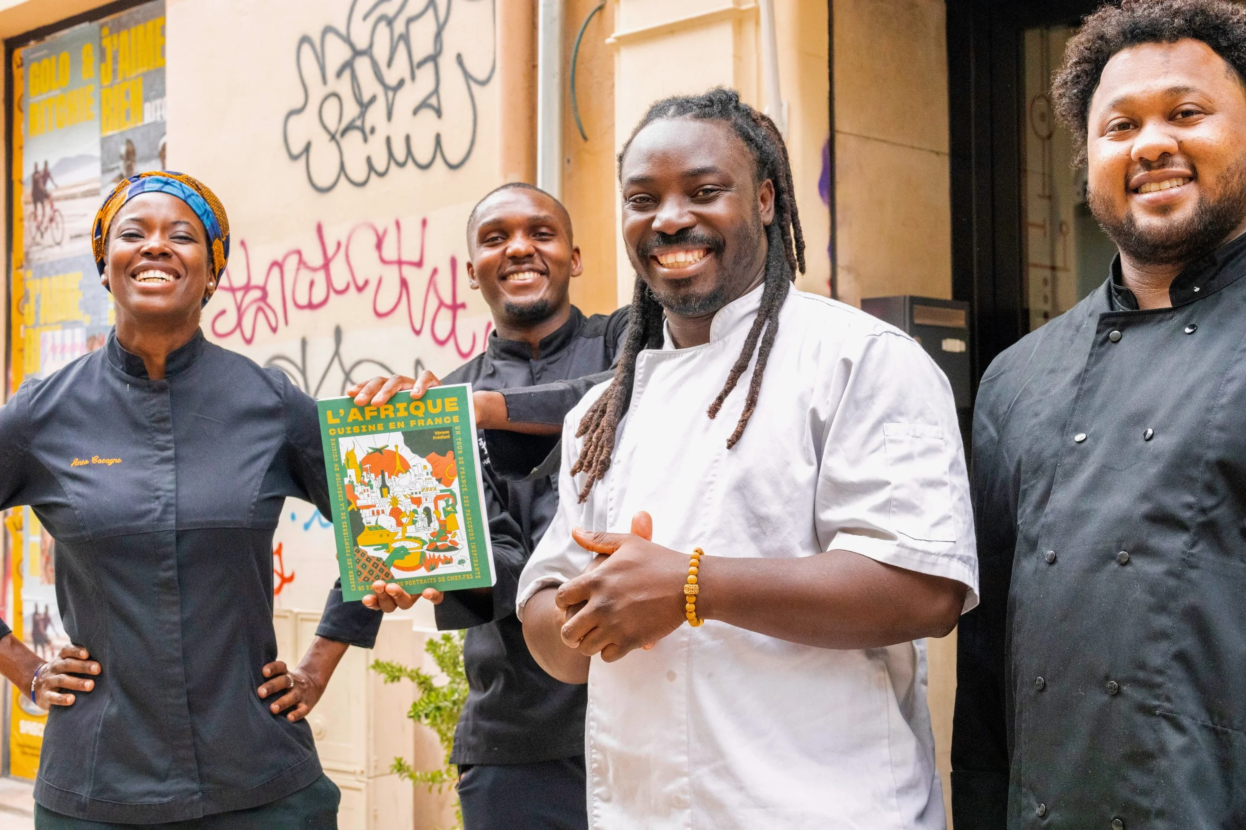 Four chefs standing together smiling, with one holding a colorful book titled "L'Afrique Cuisine en France" in front of a graffiti-covered wall.