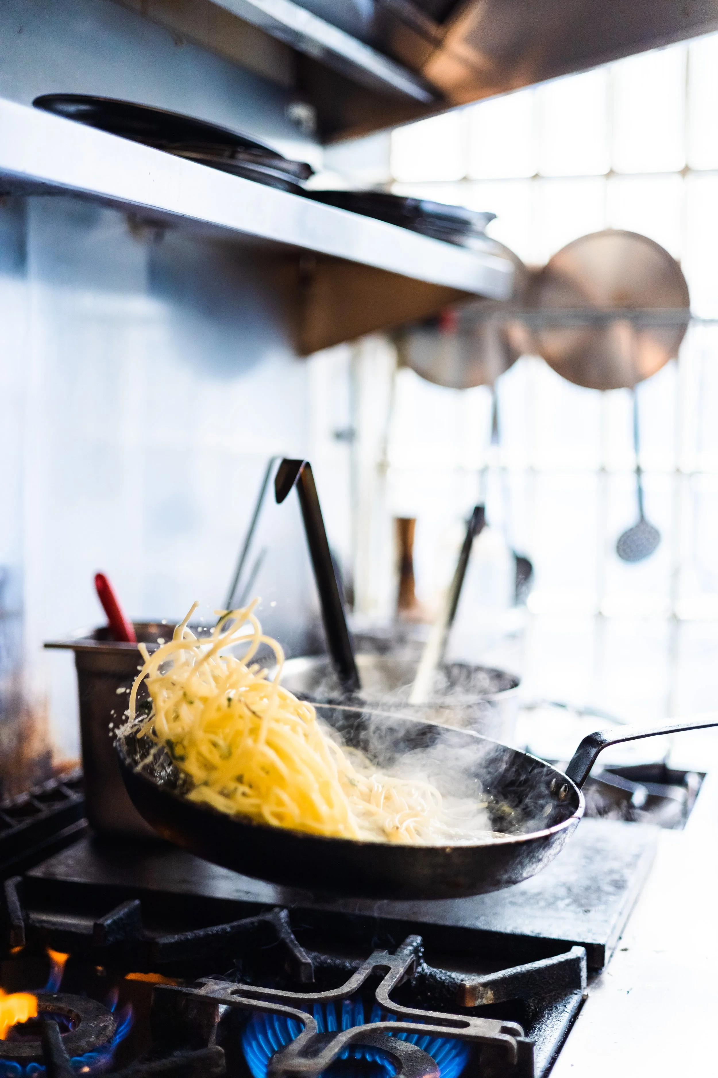 Cooking pasta in a skillet on a stovetop with steam rising, with pots and kitchen utensils in the background.