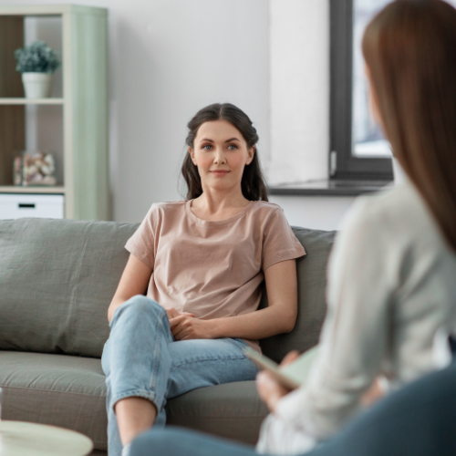 A woman sitting on a gray sofa in a therapy session, talking to a counselor in a cozy room with a white wall, window, and a shelf with decorative items.