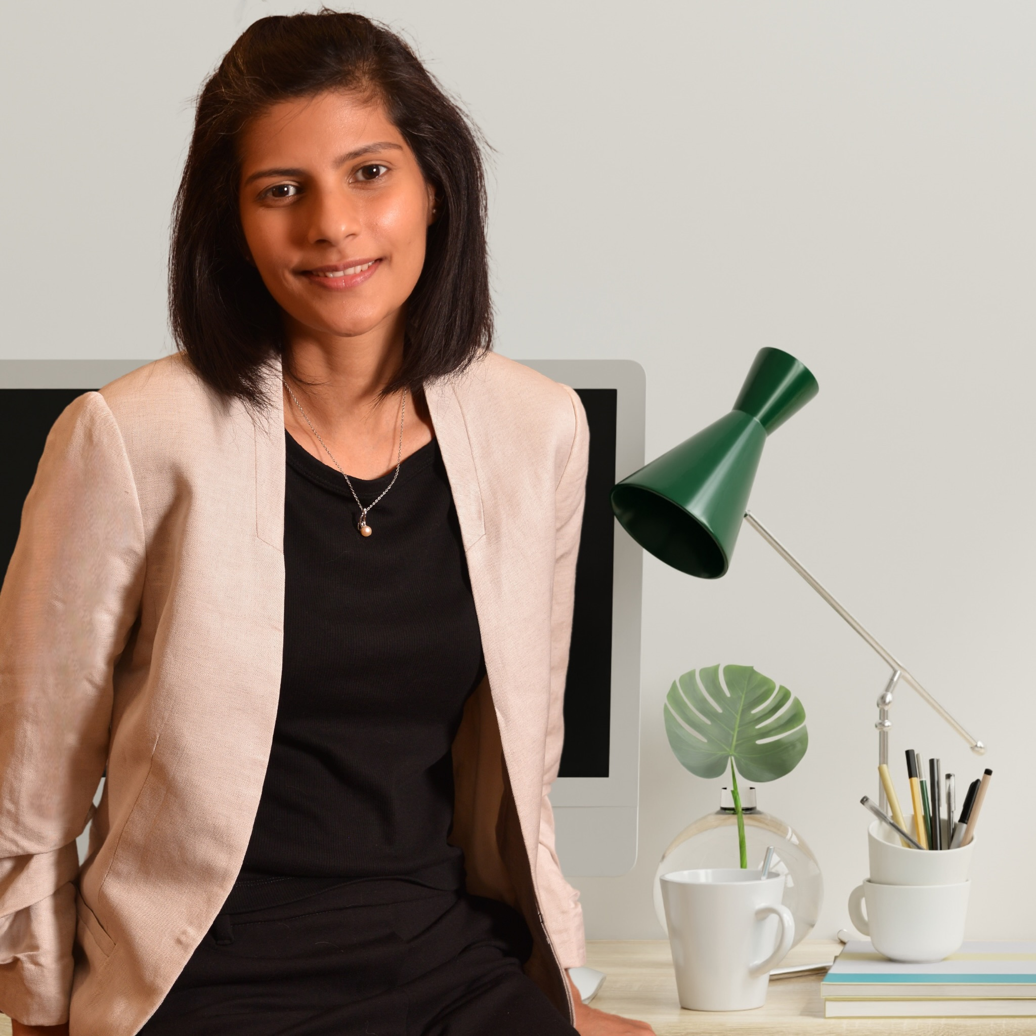 A woman sitting in a workspace with a computer, green desk lamp, a glass vase with a green leaf, and a white mug on a desk with books.