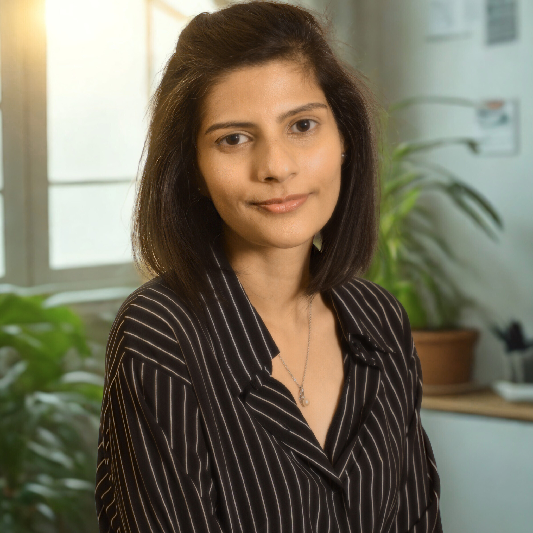 A woman with shoulder-length dark hair, wearing a black and white striped blouse and a necklace, standing in an indoor setting with plants and windows in the background.