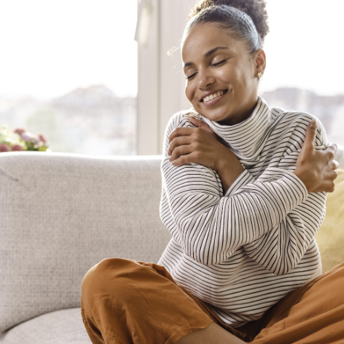Smiling woman sitting on a sofa, hugging herself with eyes closed and a content expression.
