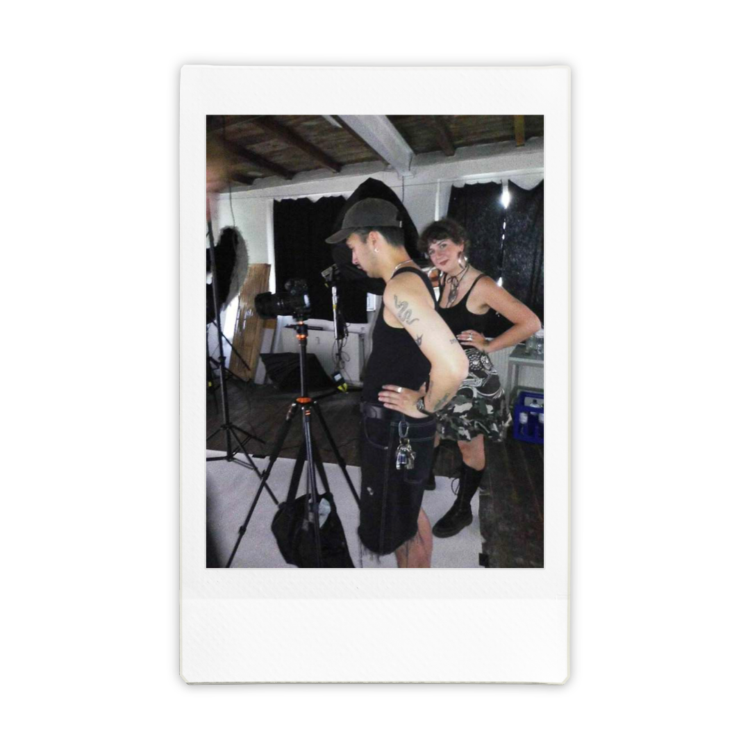 Two people working on a photoshoot: a man in black tank top and hat looking at a camera, and a woman in a black top and camouflage skirt standing with hands on hips, inside a room with black curtains and photography equipment.