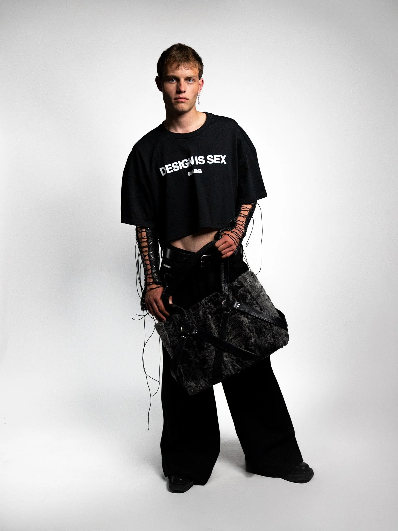 Young person with short hair wearing a black cropped T-shirt with the text 'DESIGN IS SEX' and black wide-leg pants, holding a large black and gray bag, standing against a plain light background.