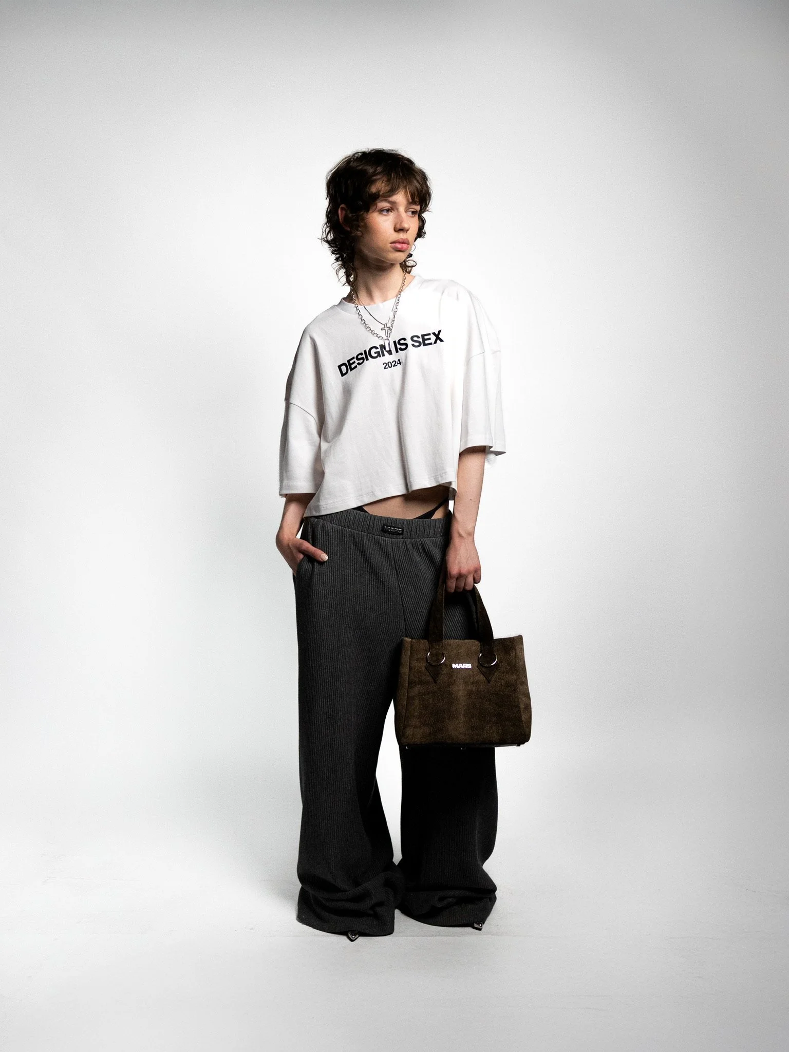 A young person with short, curly hair wearing a white T-shirt with the text "DESIGN IS SEX 2024," loose black pants, and holding a brown handbag. The person is standing against a plain white background.