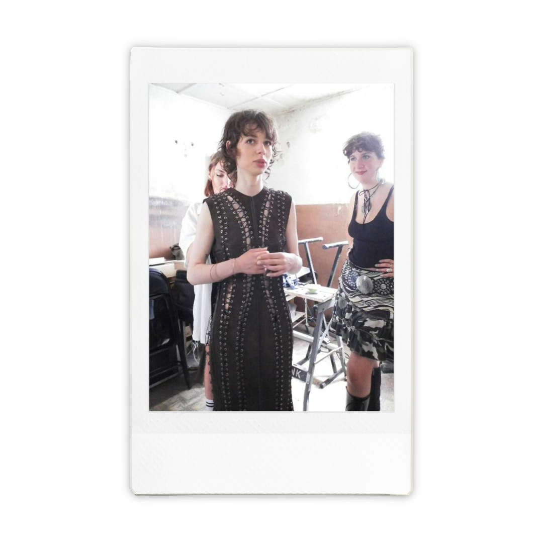Three women standing in a room, one in front wearing a black dress with metal eyelets, others in casual outfits, with a desk and chairs in the background.