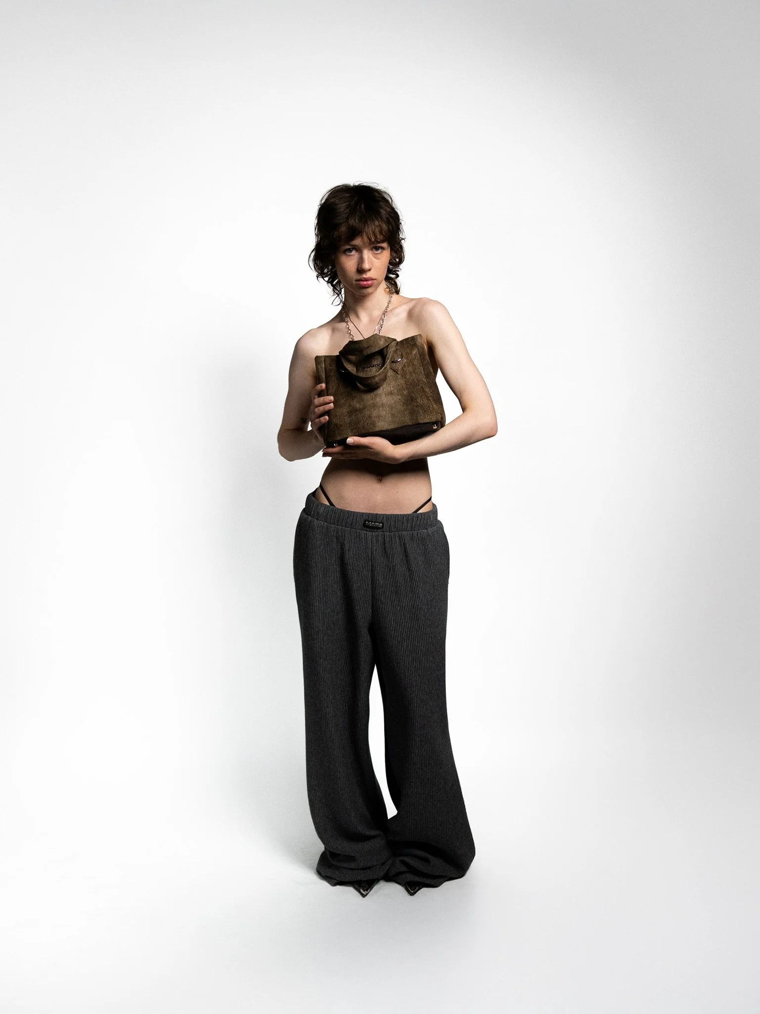 Young woman with dark hair and light skin holding a brown purse, wearing loose dark gray pants, standing against a plain white background.