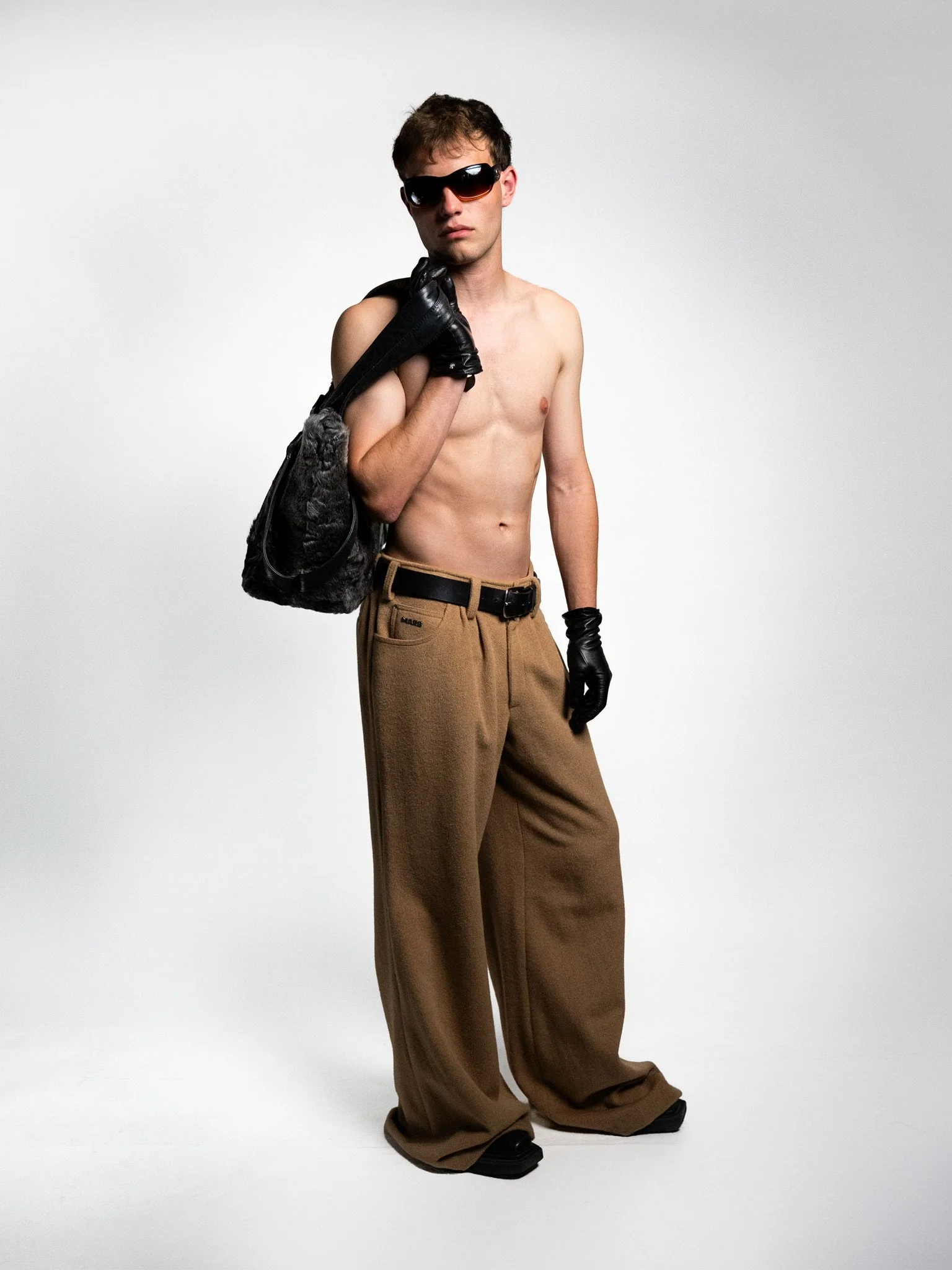 A young man with brown hair wearing sunglasses, black gloves, and holding a black bag over his shoulder, shirtless, wearing loose brown pants and black sandals, standing against a plain white background.