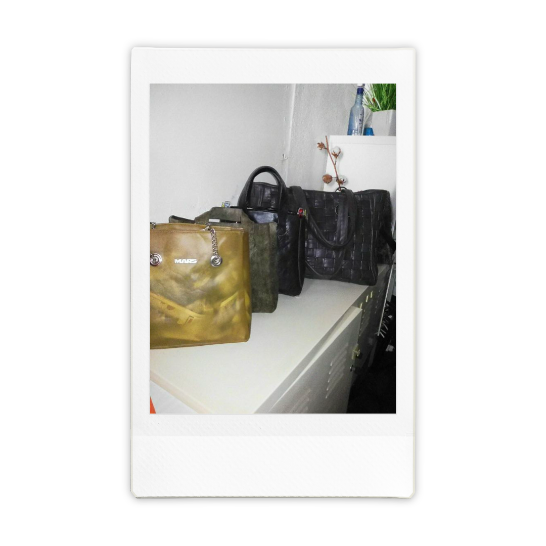 Four handbags on a white surface, with a white wall and a small shelf with a plant and bottles in the background.