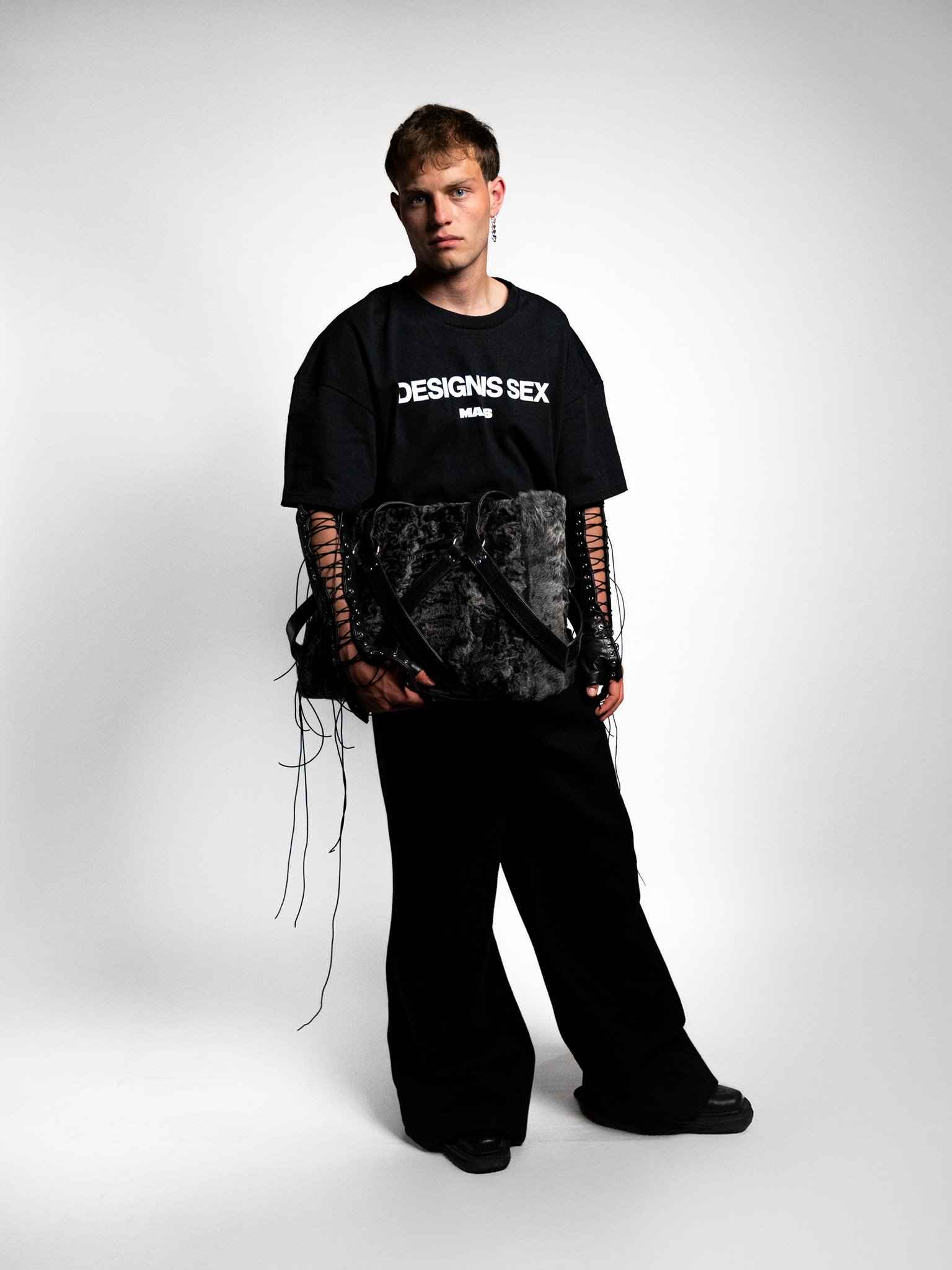 Young man wearing a black t-shirt with white text, black wide-leg pants, gloves, and holding a large fuzzy bag, standing against a plain white background.