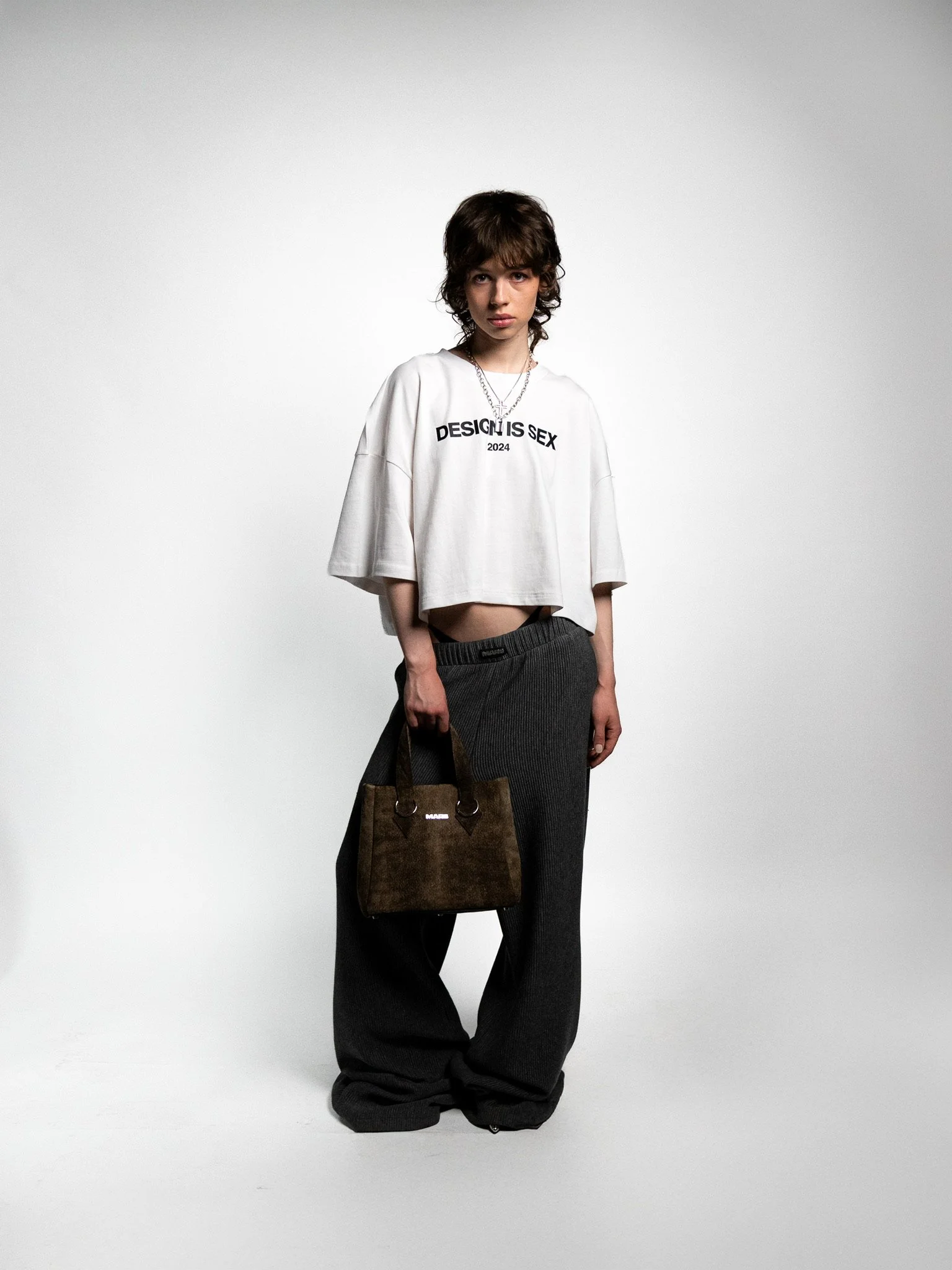 A young woman with short brown hair wearing a white cropped T-shirt with black text, loose dark gray pants, holding a brown handbag, standing against a plain gray background.
