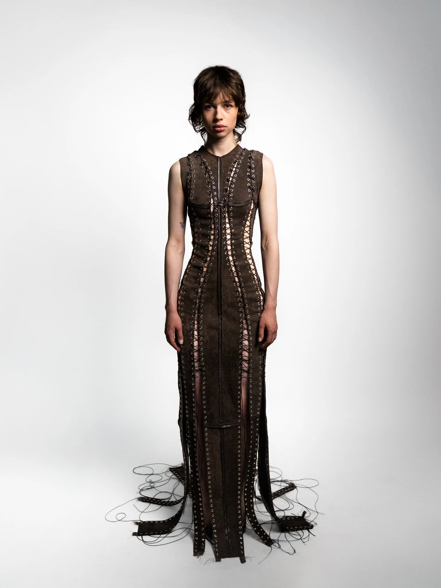 A young woman in a long, dark, sleeveless dress with metal eyelet and lace-up detailing, standing against a plain white background.