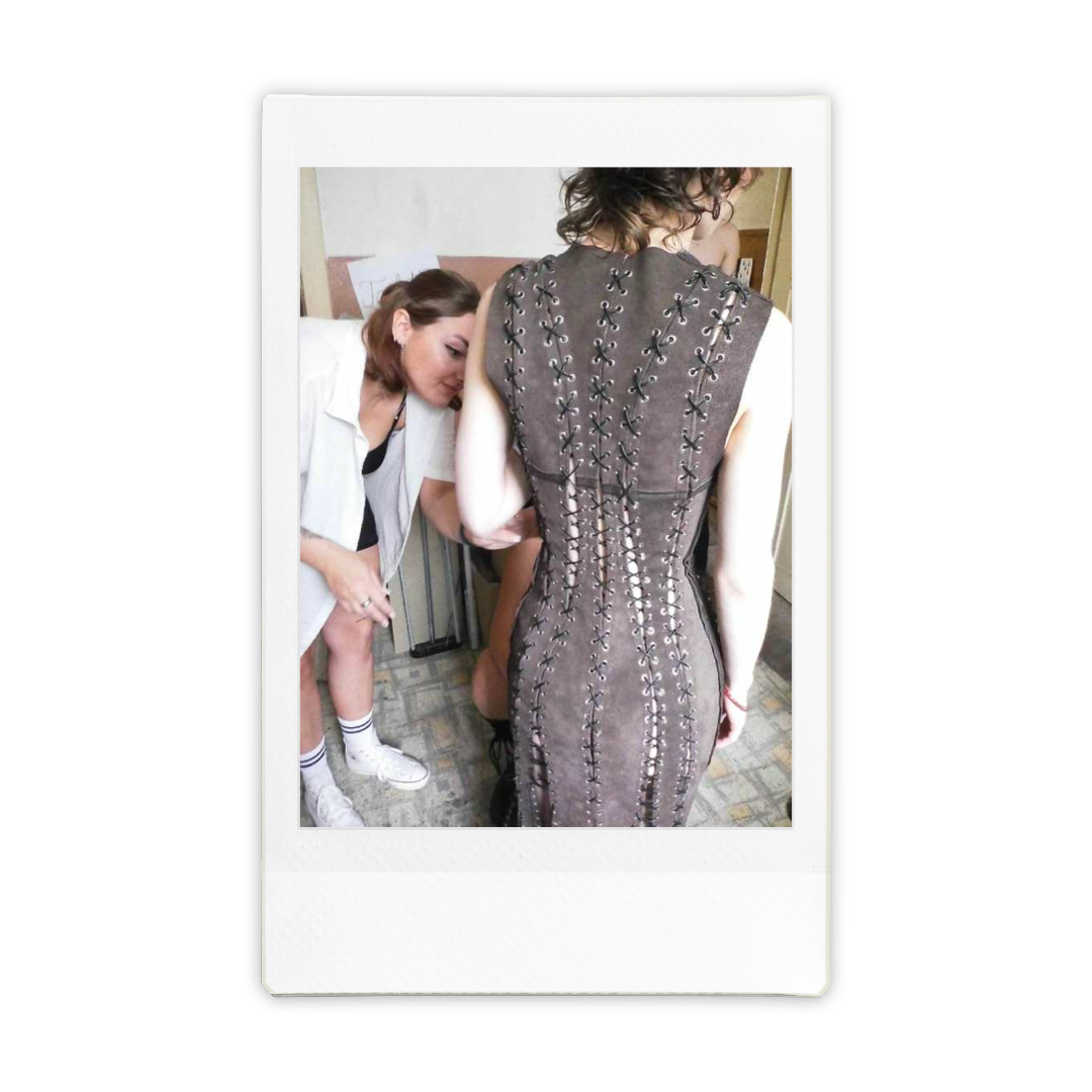 A person wearing a backless dress with lace-up detailing standing with their back to the camera, while another woman is leaning in close, observing or assisting, in a room with a patterned carpet.