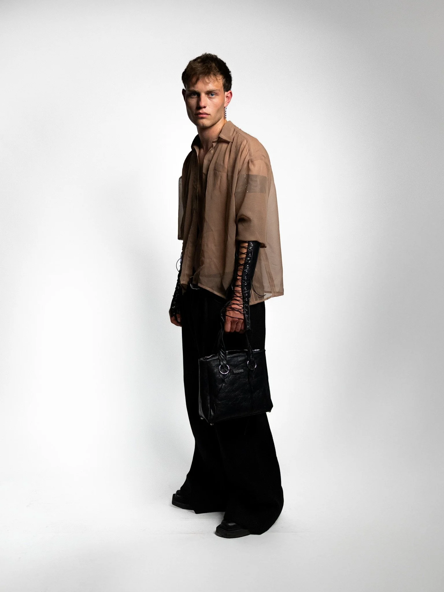 Young man wearing a sheer beige top with black lace-up gloves and black wide-leg pants, holding a black handbag, standing against a plain white background.