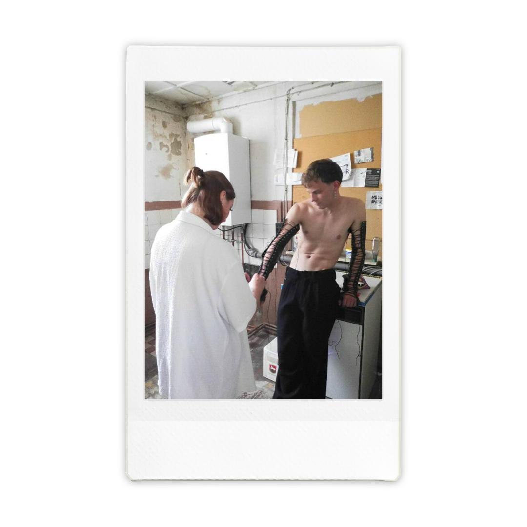 A young man with no shirt and black pants is receiving a test or treatment involving long black tubes on his arms from a woman in a white coat in a clinical setting.