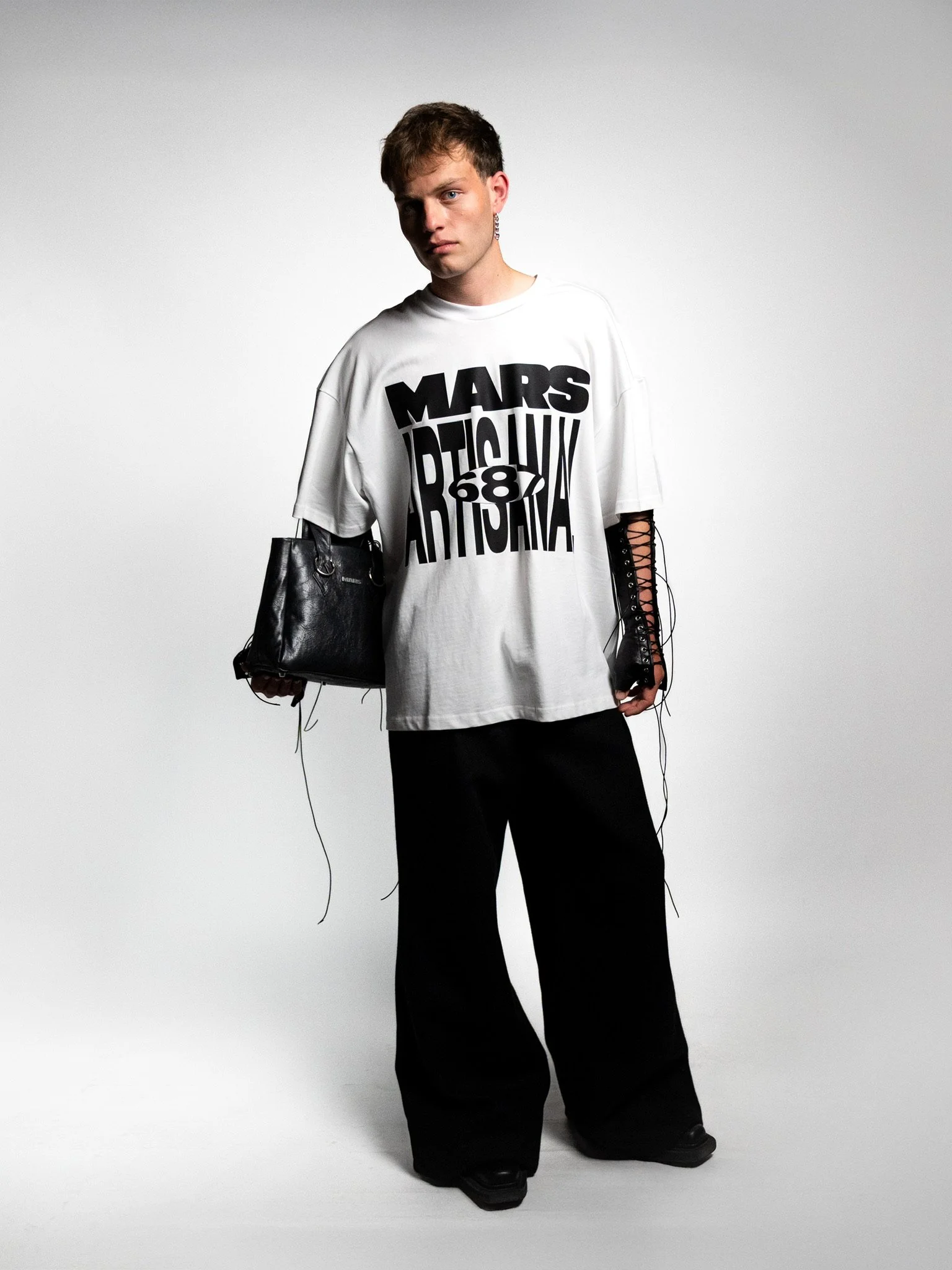 A young man with short, light brown hair wearing a white oversized T-shirt with black graphic text and numbers, black wide-leg pants, and black shoes stands against a plain light gray background. He carries a black handbag on his left arm and has bla