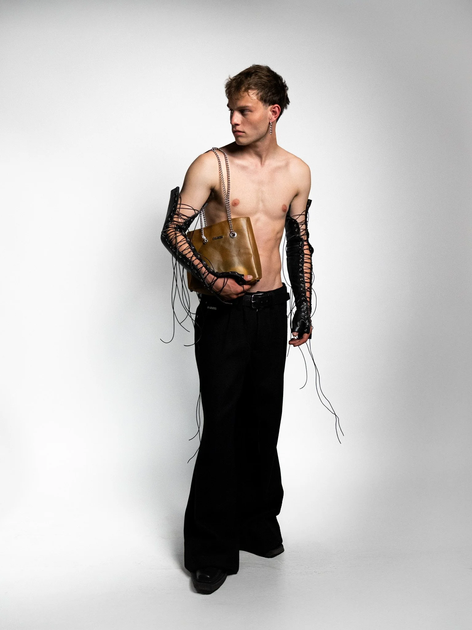 A young man standing shirtless against a plain background, holding a brown handbag, wearing black wide-leg pants, and long black gloves with lacing details.