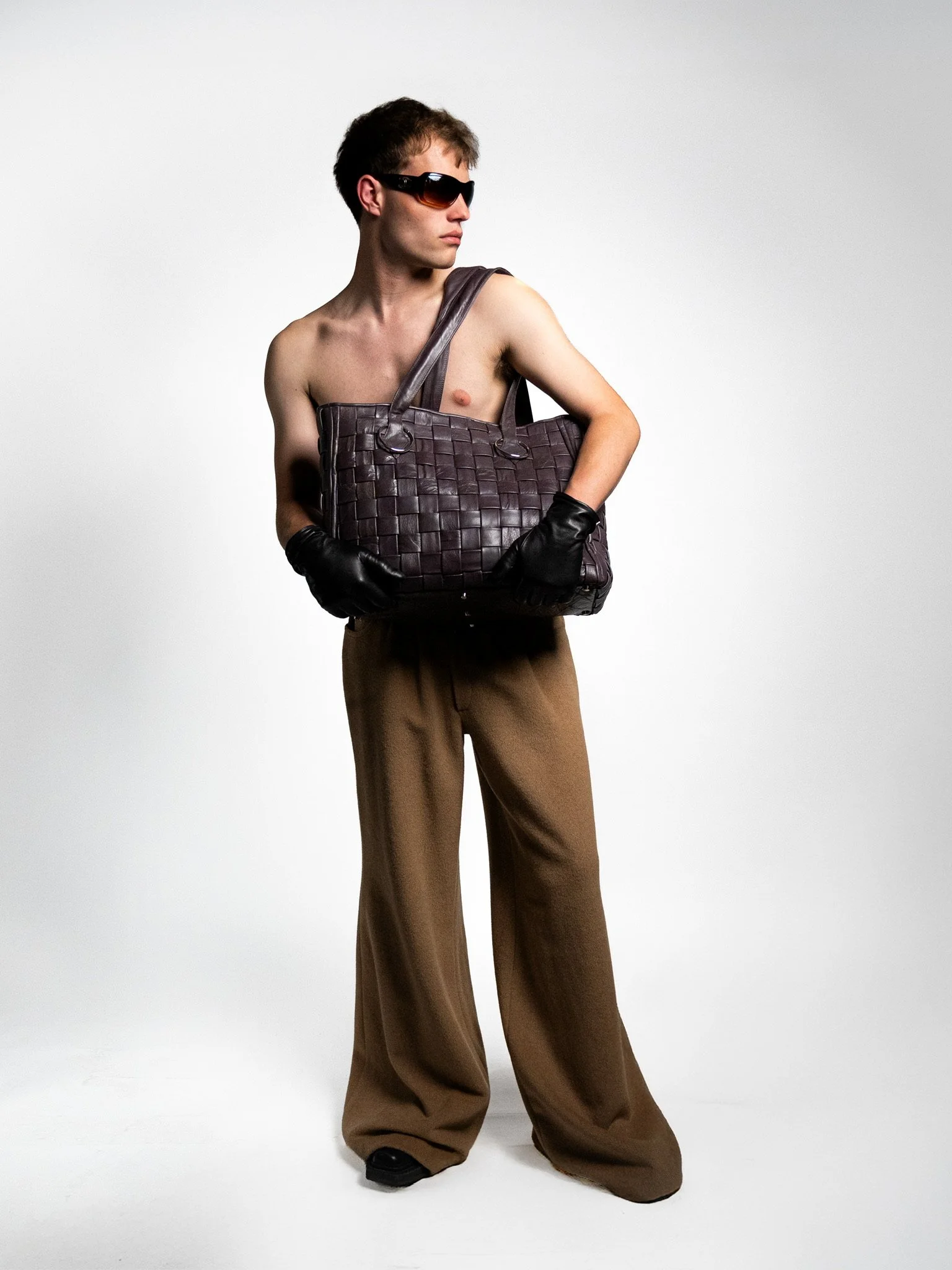 Young man with sunglasses, shirtless, wearing brown wide-leg pants, black gloves, and holding a large woven leather bag, standing against a plain white background.