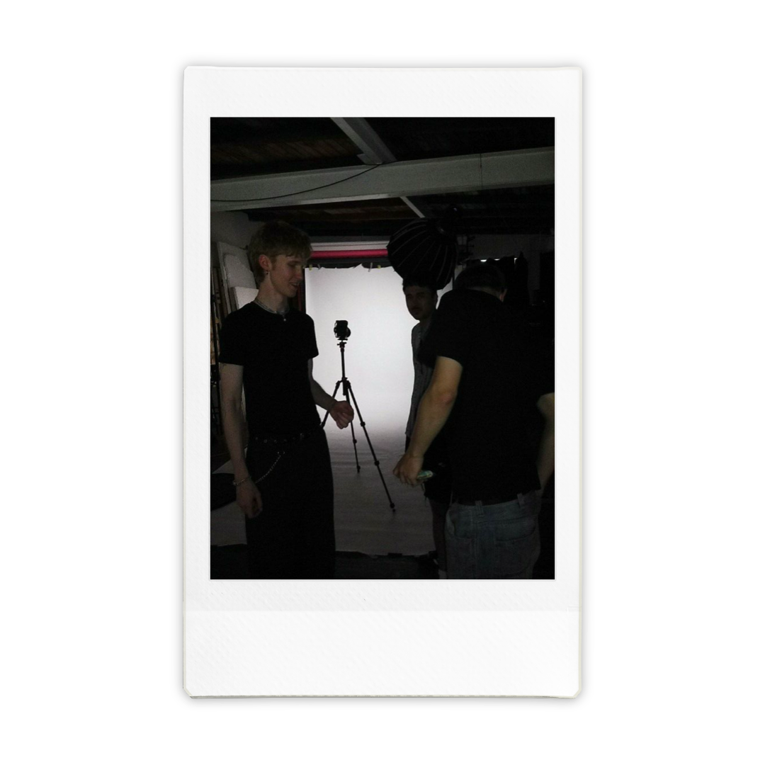 Three young men in a photography studio with a camera on a tripod and a white backdrop, seemingly preparing for a photoshoot.