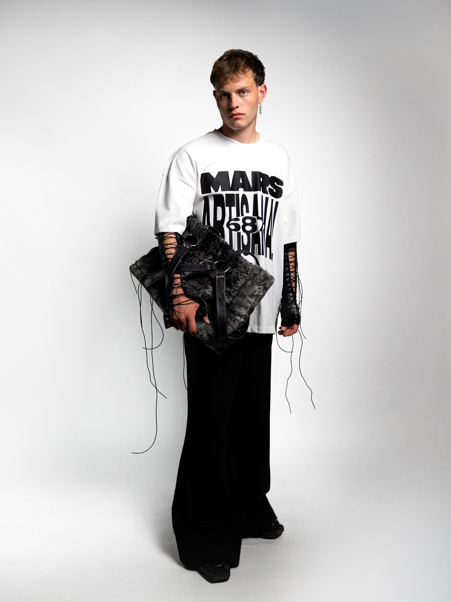 A young person with short hair and earrings standing against a plain background, wearing a white oversized t-shirt with black bold text, black wide-leg pants, and holding a large black bag with decorative strings.