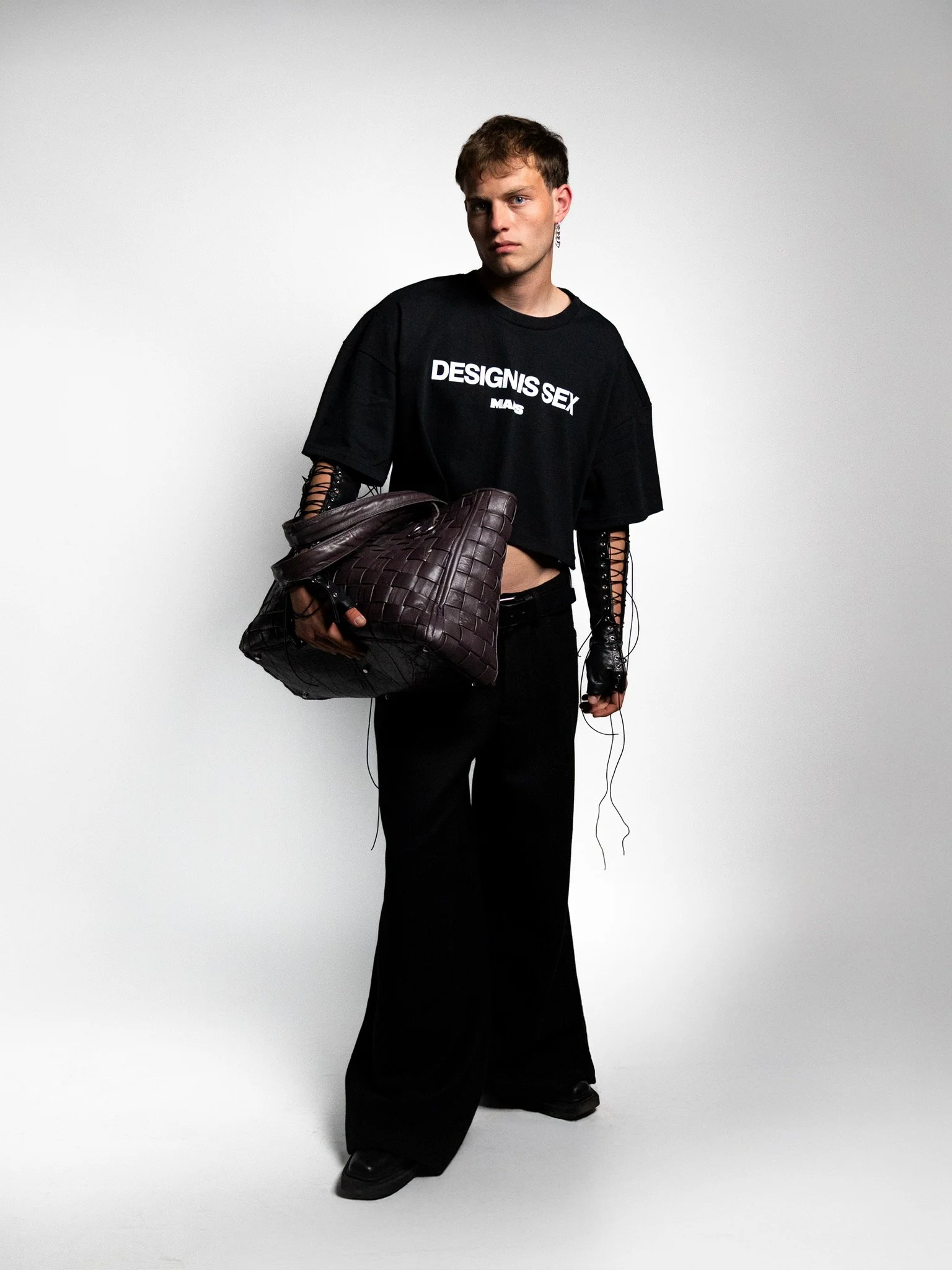 A person standing against a plain white background, wearing a black cropped T-shirt with white text, black wide-leg pants, gloves, and holding a large woven dark bag.