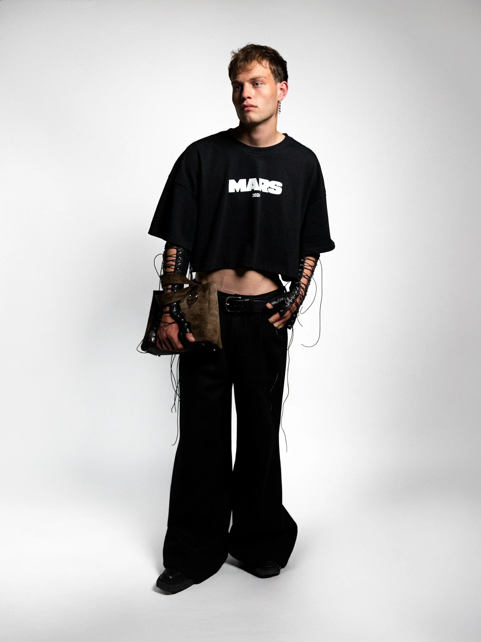 A young man with short, light brown hair posing against a plain gray background. He is wearing a black, cropped t-shirt with 'MARS 2021' printed on it, loose black pants, and black shoes. He has black gloves with lace-up sleeves that extend from his 