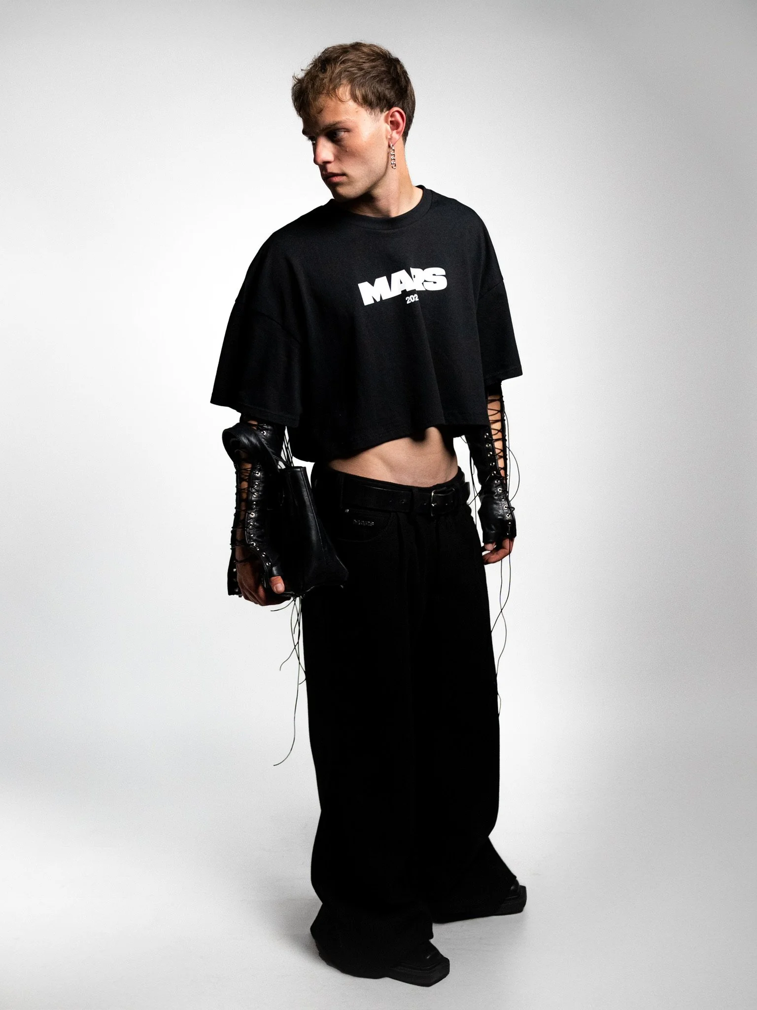 A young man with light brown hair and earrings wearing a black crop top T-shirt with white text, black baggy pants, black fingerless gloves, and carrying a black bag, standing against a plain gray background.