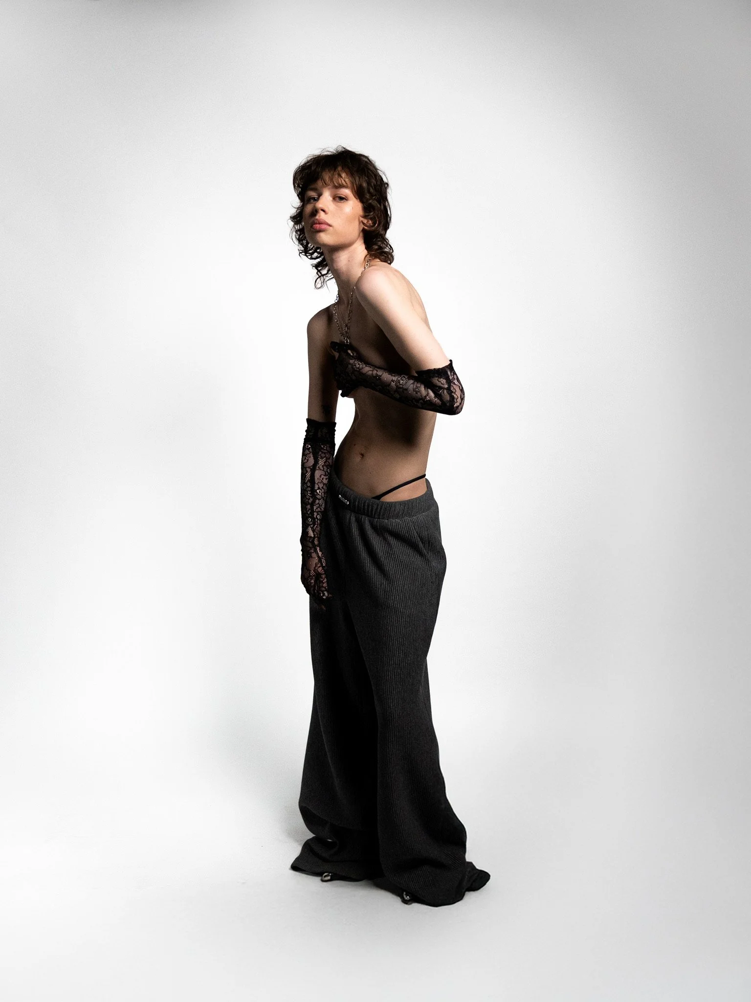 A young person with dark curly hair, wearing long lace gloves, underwear, and loose dark pants, standing against a plain light background.