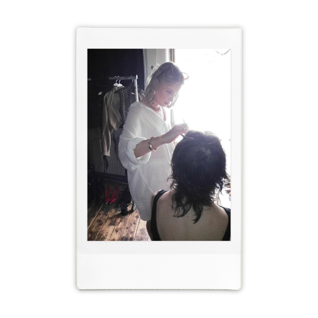 A woman sitting with her back to the camera is having her hair styled by a hairdresser who is standing, with sunlight coming through a window behind them.