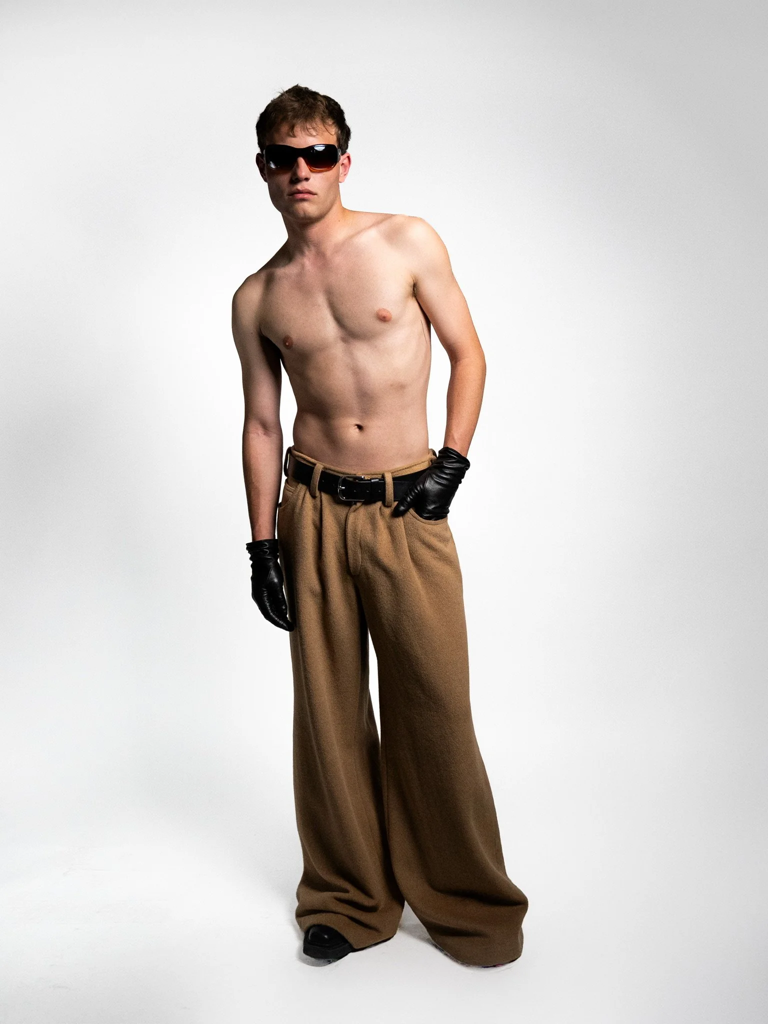 A shirtless young man wearing large sunglasses, black gloves, loose brown pants, and black shoes, standing against a light gray background.