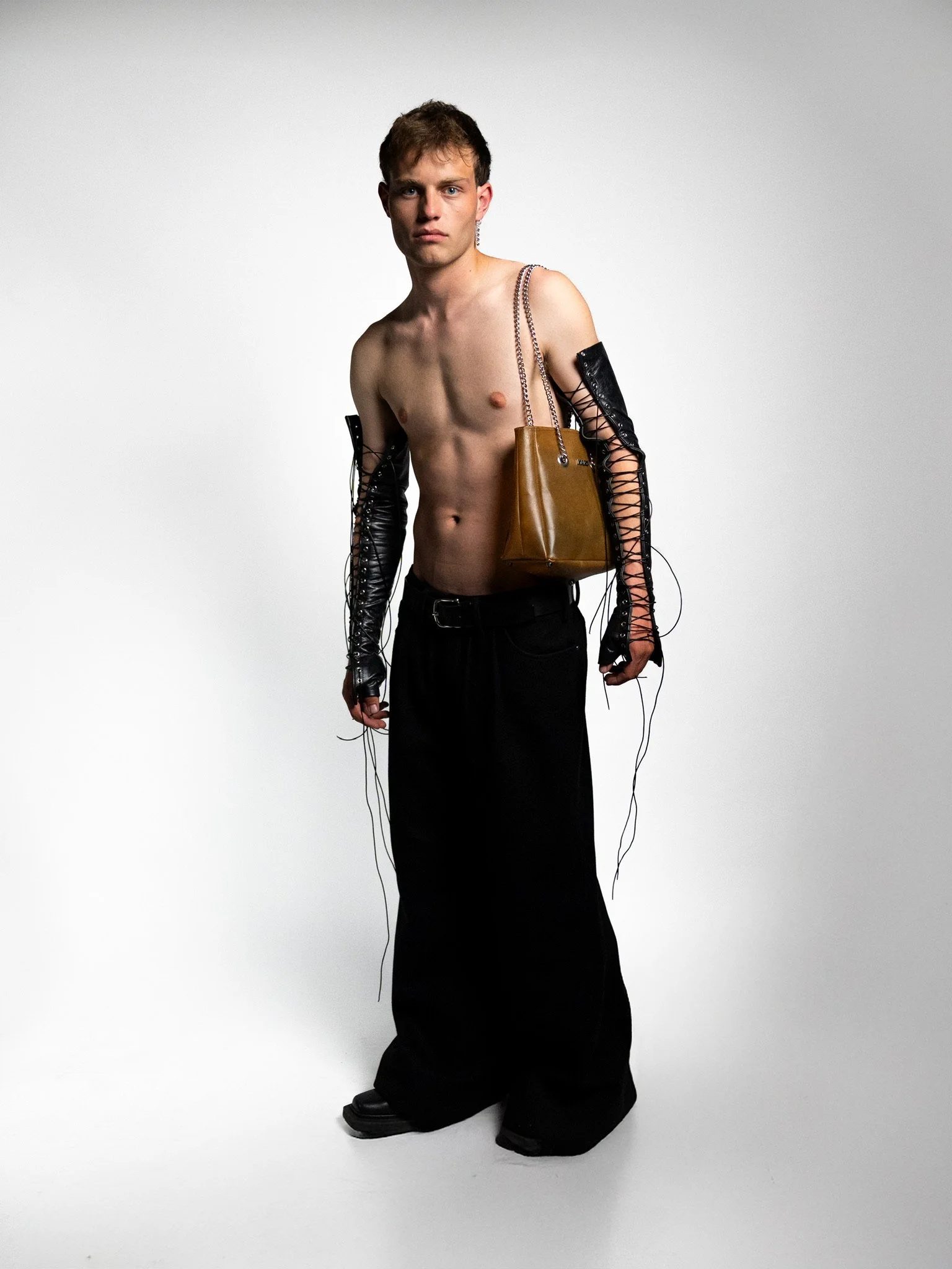 A young man with short brown hair, shirtless, wearing black wide-legged pants, black lace-up arm sleeves, and carrying a brown handbag over his shoulder, standing against a plain gray background.