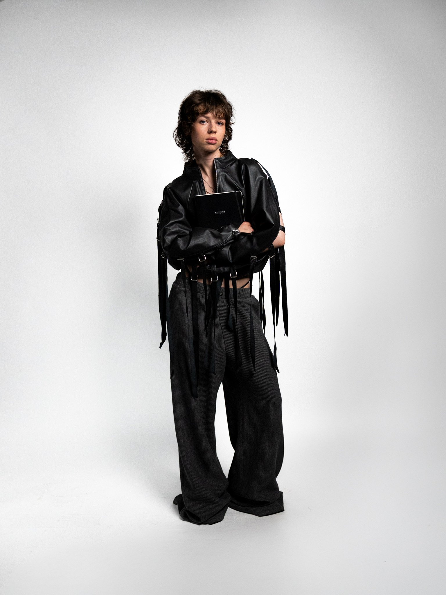 A young person with short, wavy brown hair, dressed in a black leather jacket, dark gray wide-leg pants, holding a small black notebook, standing against a plain white background.
