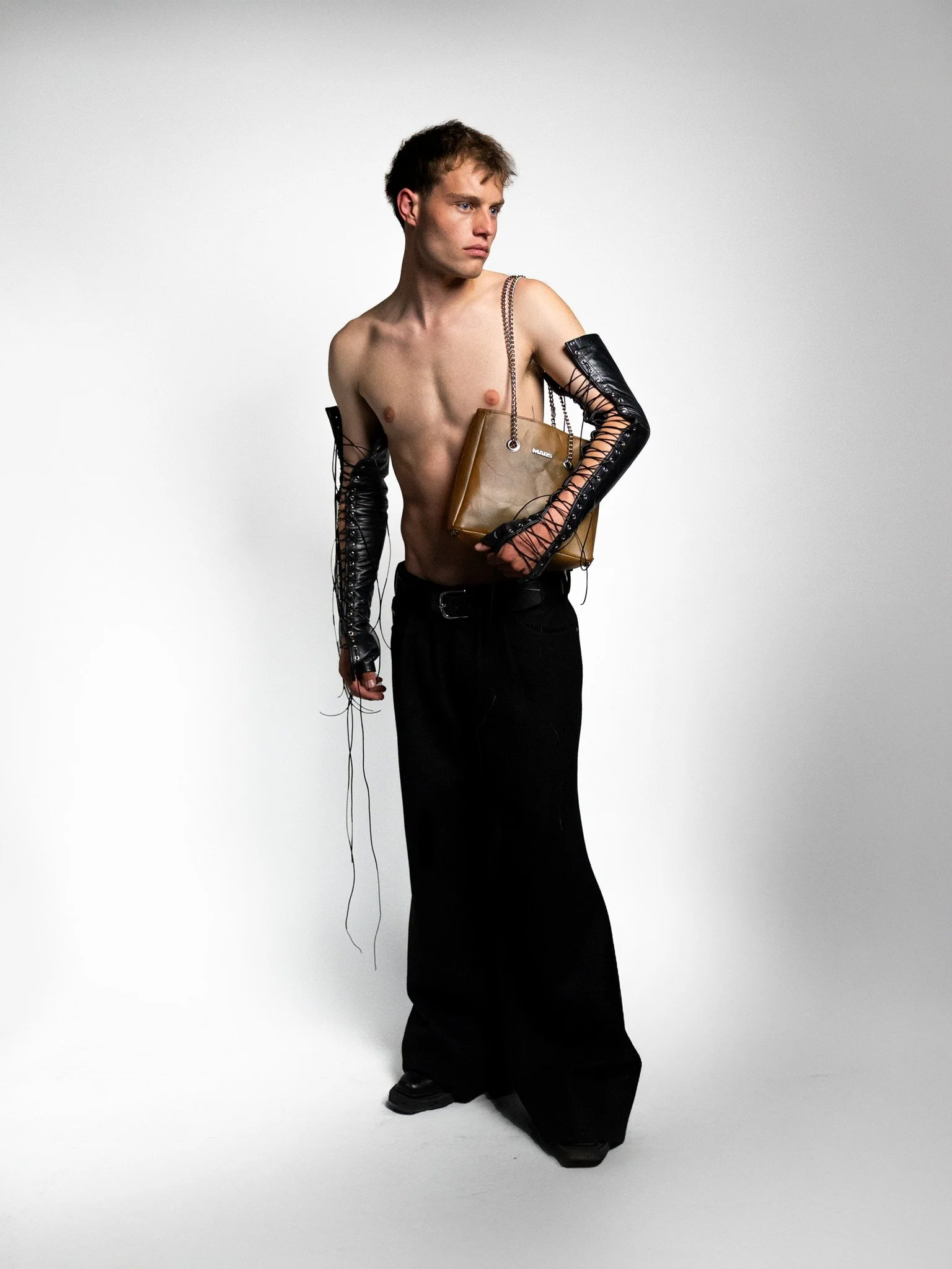 A shirtless young man with black wide-leg pants and long plastic arms with laces holds a brown handbag and looks to the side against a plain light gray background.