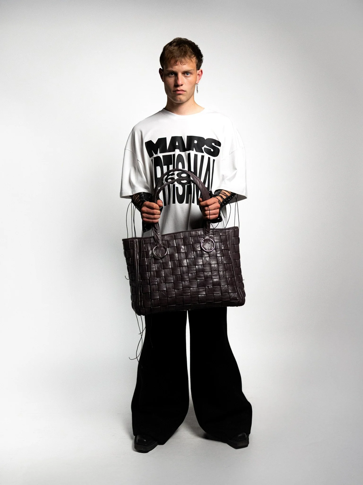 A young person with short brown hair wearing a white oversized T-shirt with black text, black wide-leg pants, and holding a large woven dark purple handbag, standing against a plain light gray background.