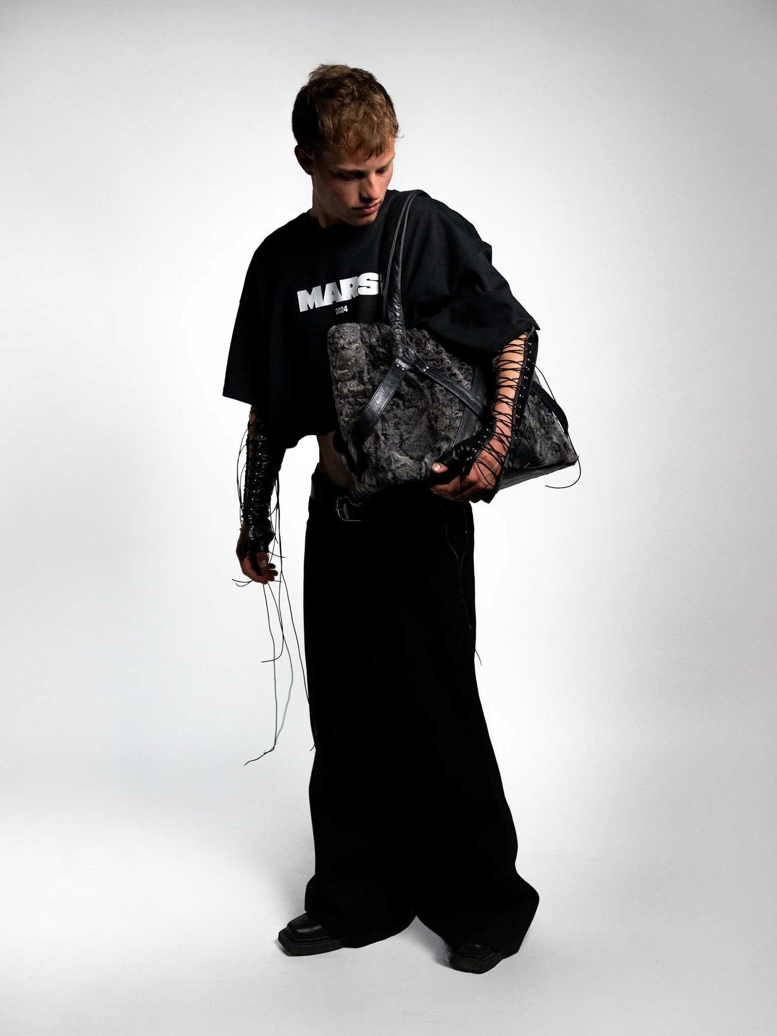 A person standing against a plain background, wearing a black oversized t-shirt with white lettering, black wide-leg pants, black shoes, and holding a large gray fur-patterned bag, with long lace-up sleeves on the t-shirt.