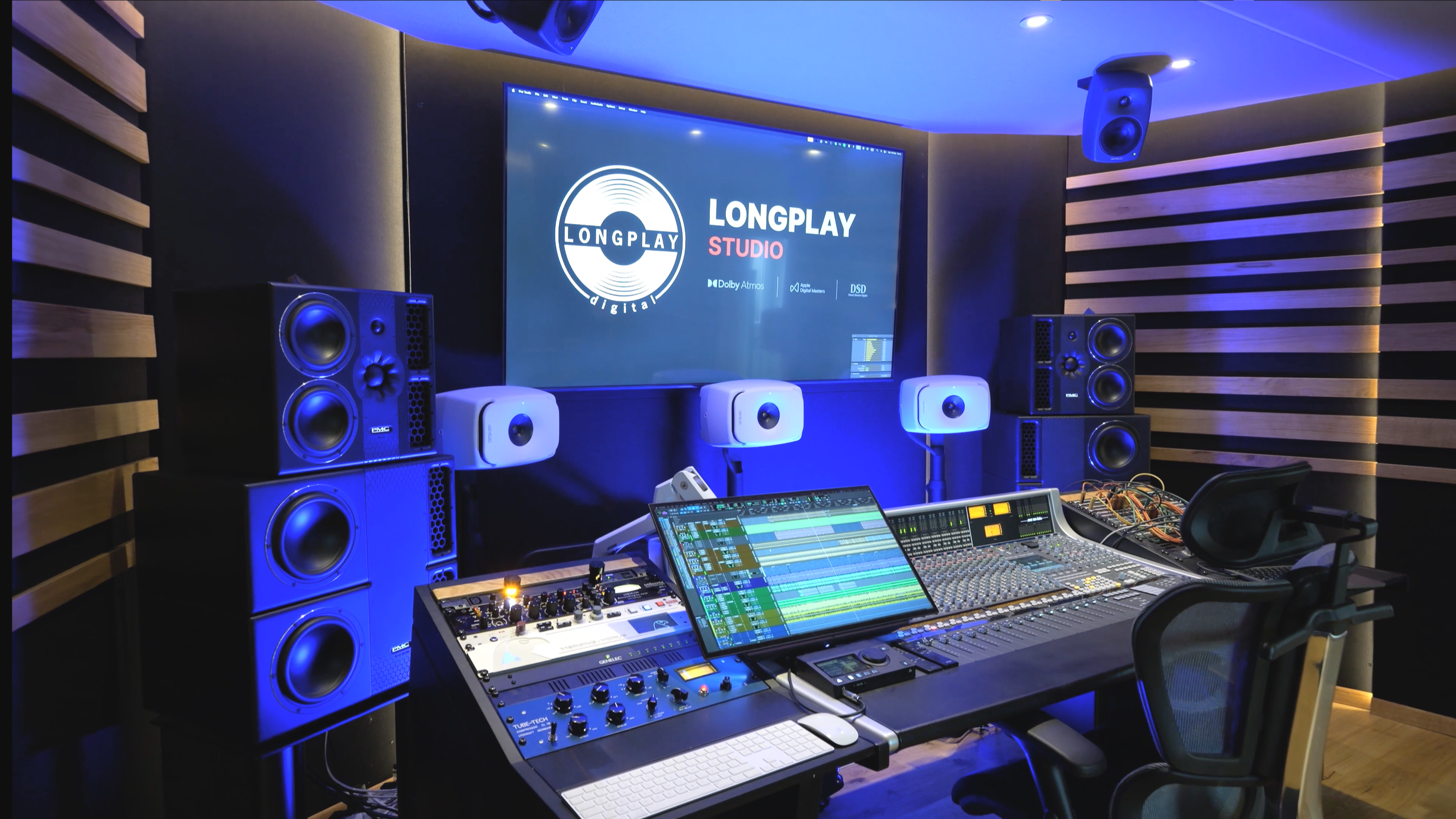Recording studio with a mixing console, computer screens displaying music software, large speakers, and acoustic paneling.