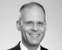 Black-and-white photo of a smiling man with glasses and receding hair, wearing a collared shirt.