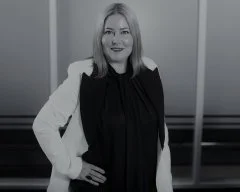 A woman smiling, standing with her hand on her hip, wearing a black top and a light-colored blazer in a modern indoor setting.